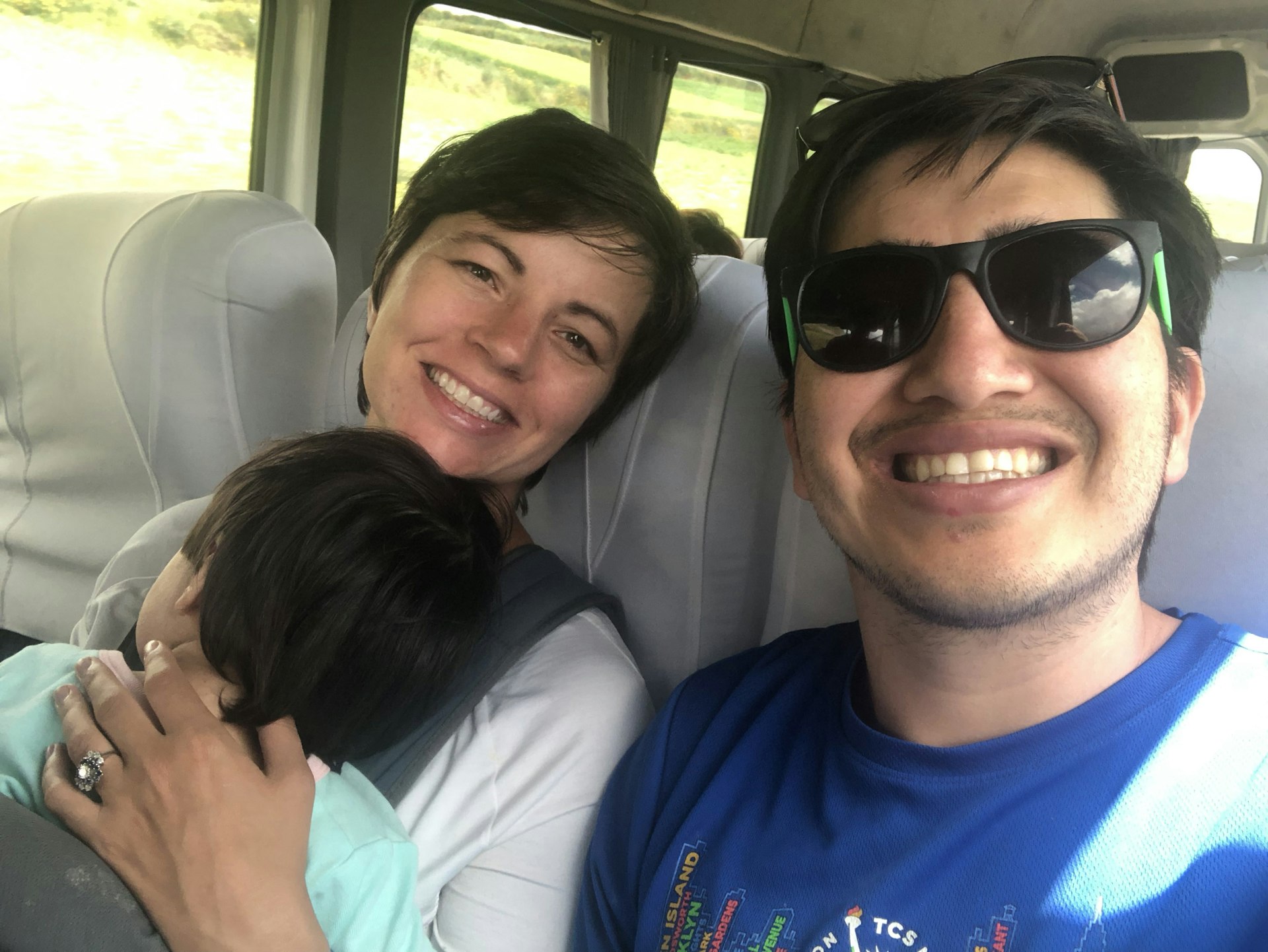 A woman cradles a sleeping baby as she smiles at the camera with a man wearing sunglasses on a train.