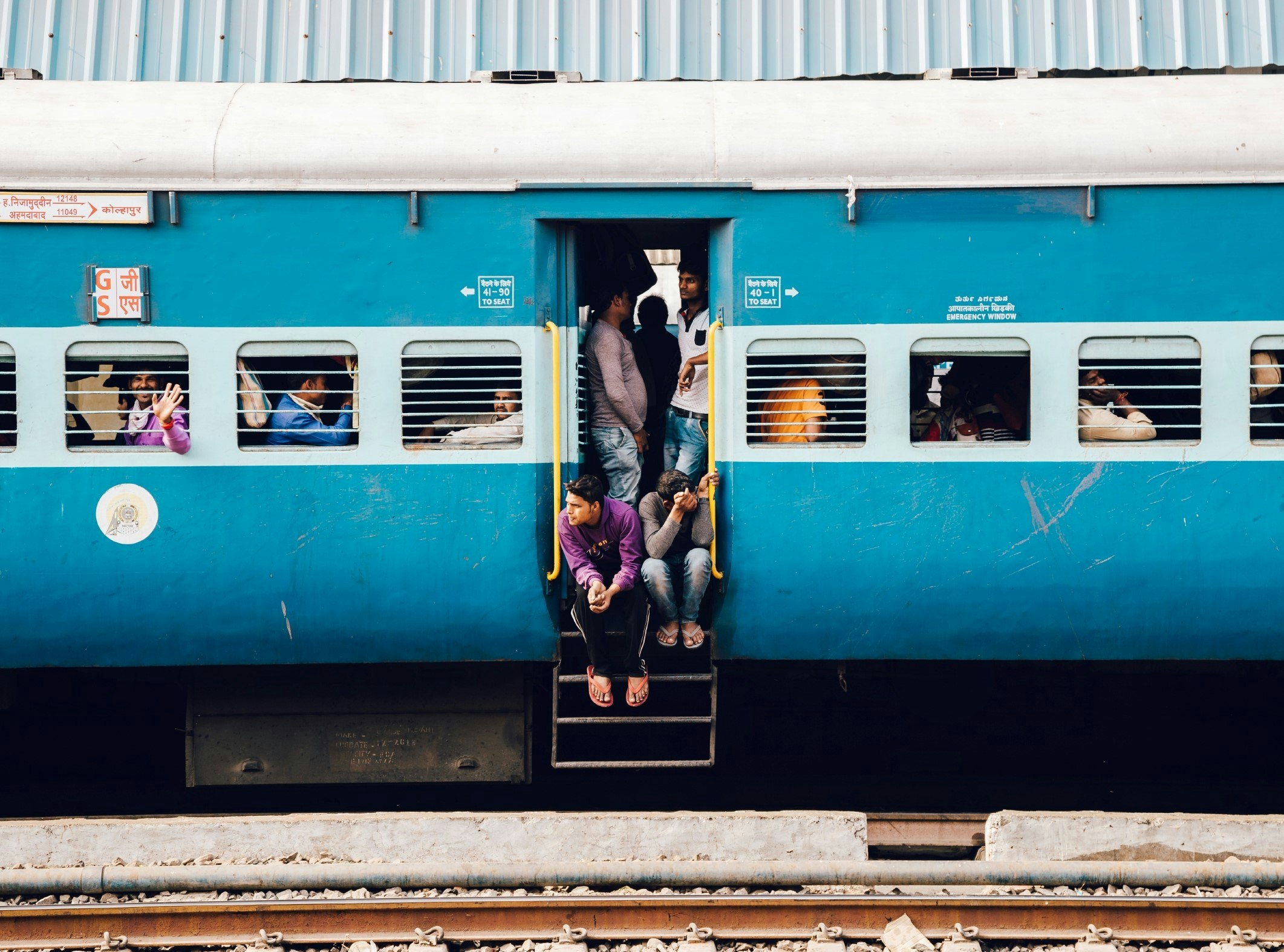 A stationary blue train in India, with passengers sitting and leaning out of the doorway and looking through the train's windows.