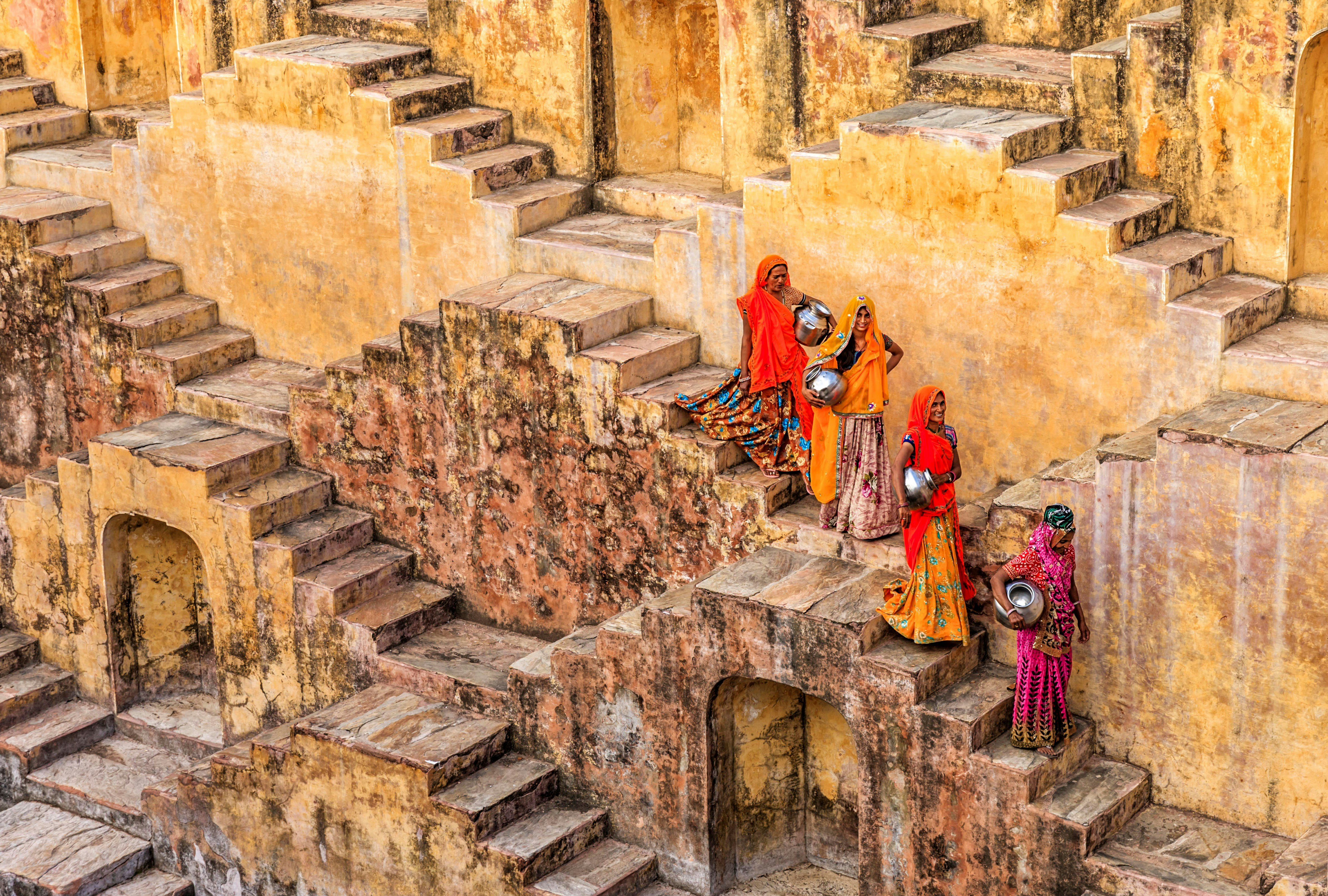 A posed picture of four women, carrying metallic pots, walking down the stairs of a stone stepwell in Rajasthan, India. The four women wear colourful saris that stand out against the stone background.
