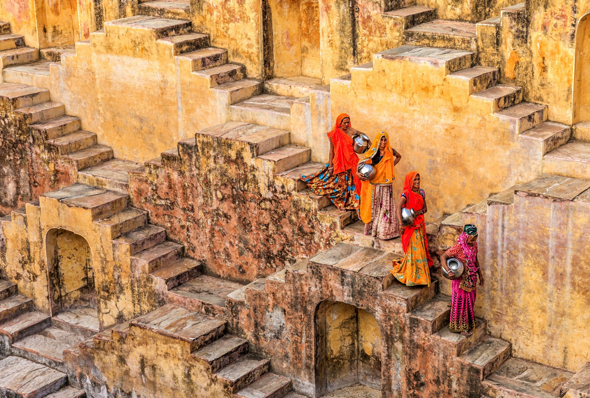 A posed picture of four women, carrying metallic pots, walking down the stairs of a stone stepwell in Rajasthan, India. The four women wear colourful saris that stand out against the stone background.