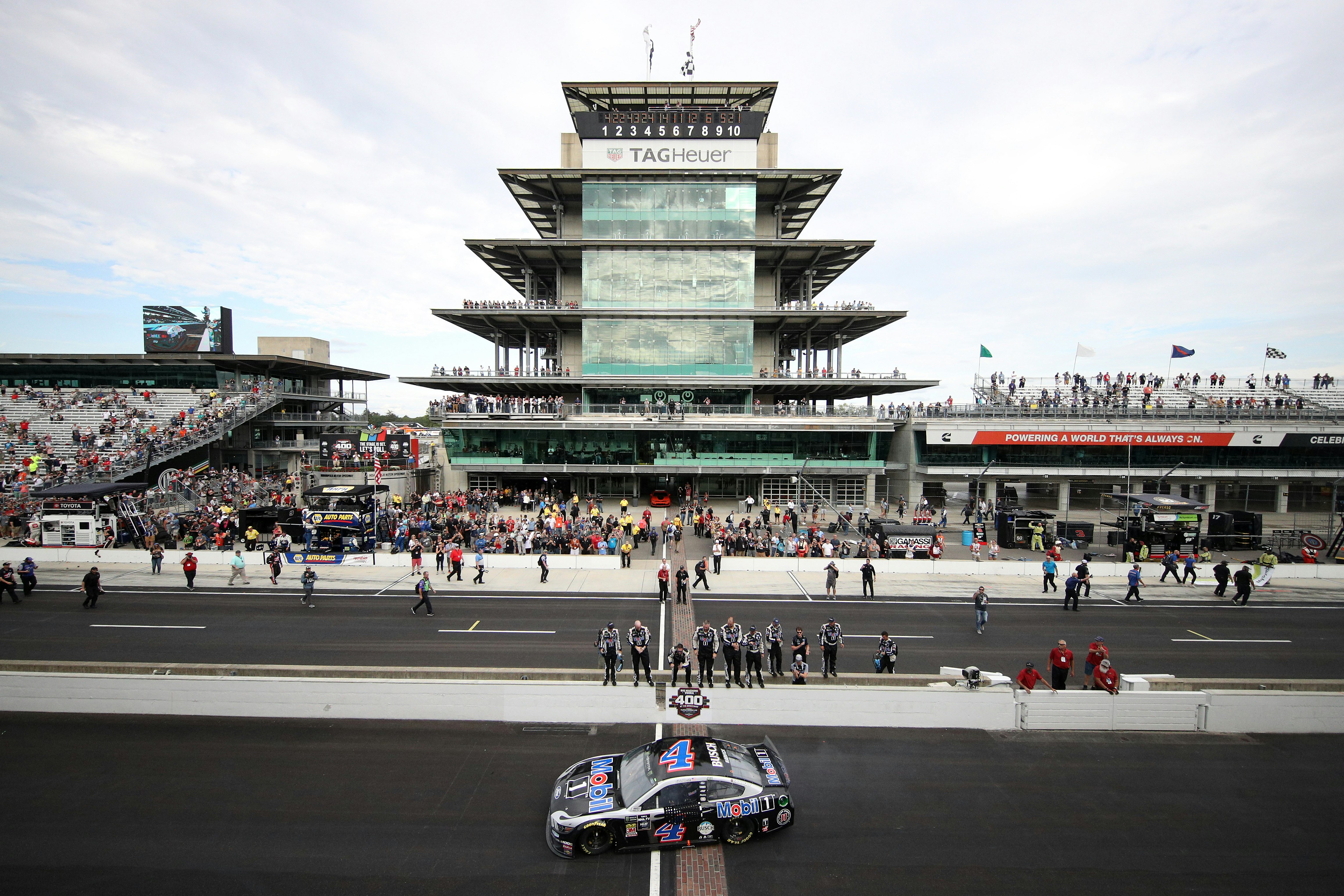 A race car drives over the iconic brick lane at the Indianapolis Motor Speedway. In the background, a large crowd is sitting in the stands.