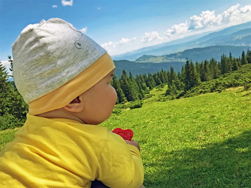 A very young baby wears bright yellow pajamas and a knit hat as he looks out on a mountain vista; kids outdoors adventures