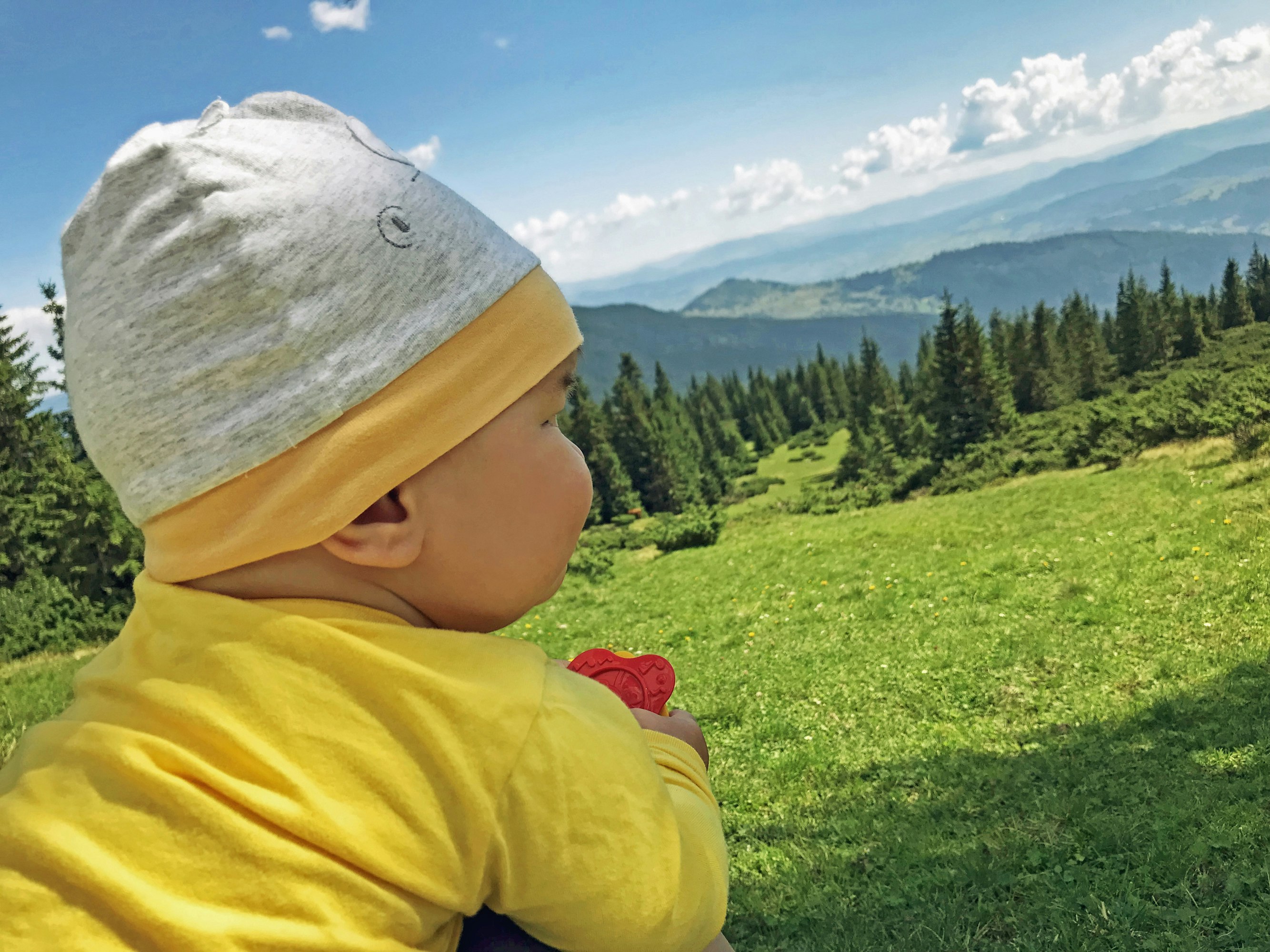 A very young baby wears bright yellow pajamas and a knit hat as he looks out on a mountain vista; kids outdoors adventures