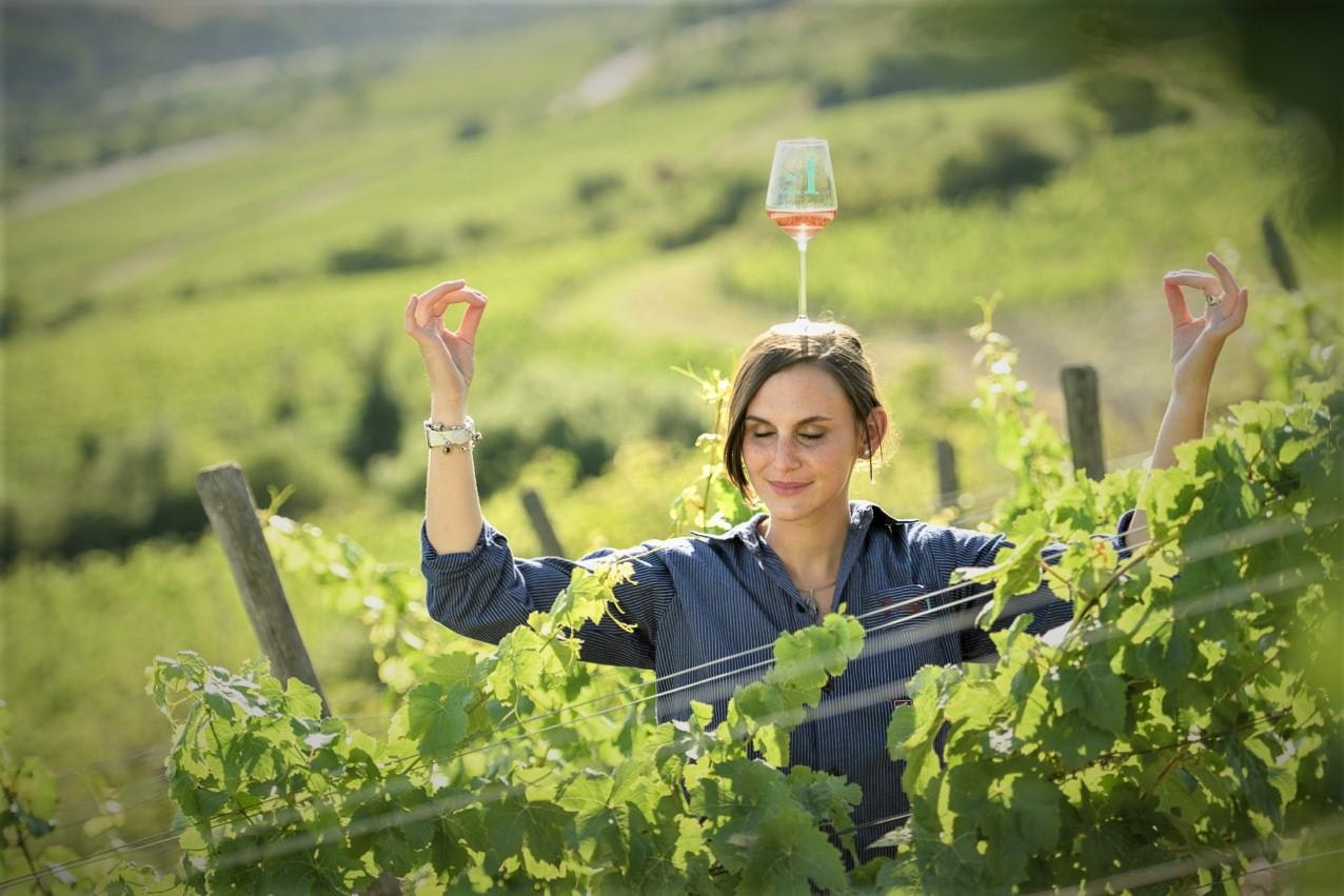 German Wine Princess Inga Storck stands in a vineyard at Schwarzer Herrgott surrounded by green leafy gapevines. She wears a blue pinstripe button-down shirt and has on a silver necklace. She is balancing a glass of rose on her head with her fingers in a meditative mudra pose and a serene look on her face, with a Mona Lisa smile