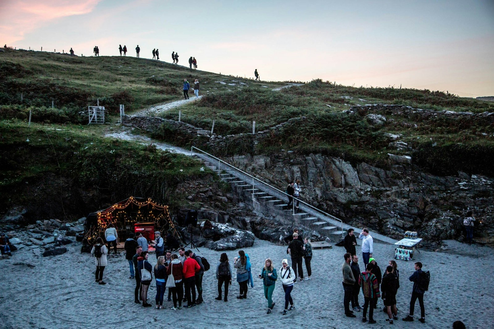 Young people standing on the beach as the sun is going down. There is a small stage surrounded by fairy lights on the beach