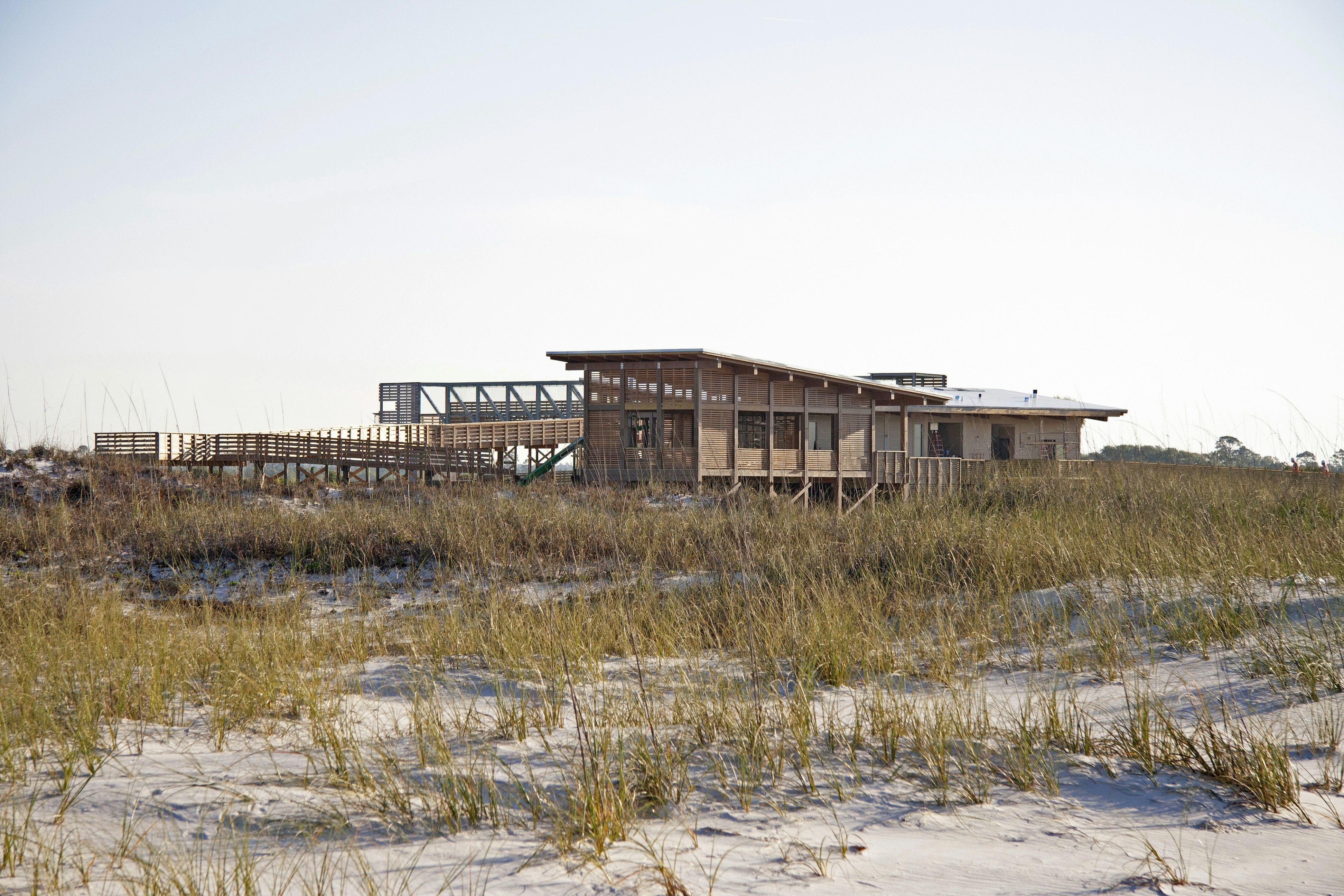 A large, modern wooden structure sits amidst sand and patches of coastal grasses. 