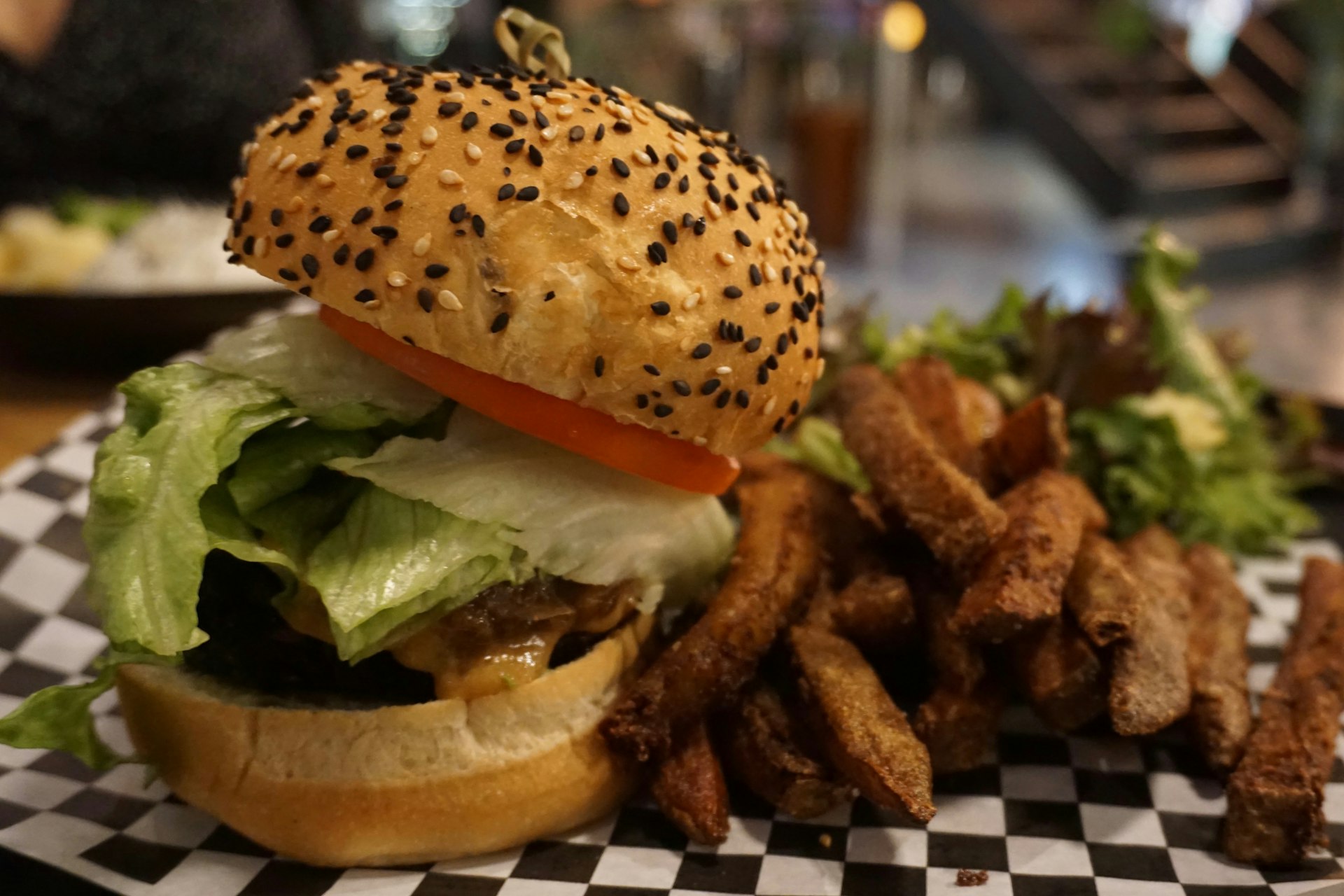 A large vegan burger is sitting on a piece of checkerboard-print paper alongside well-done fries and salad leaves. The burger is stuffed with salad and the bun is topped with black and white sesame seeds.