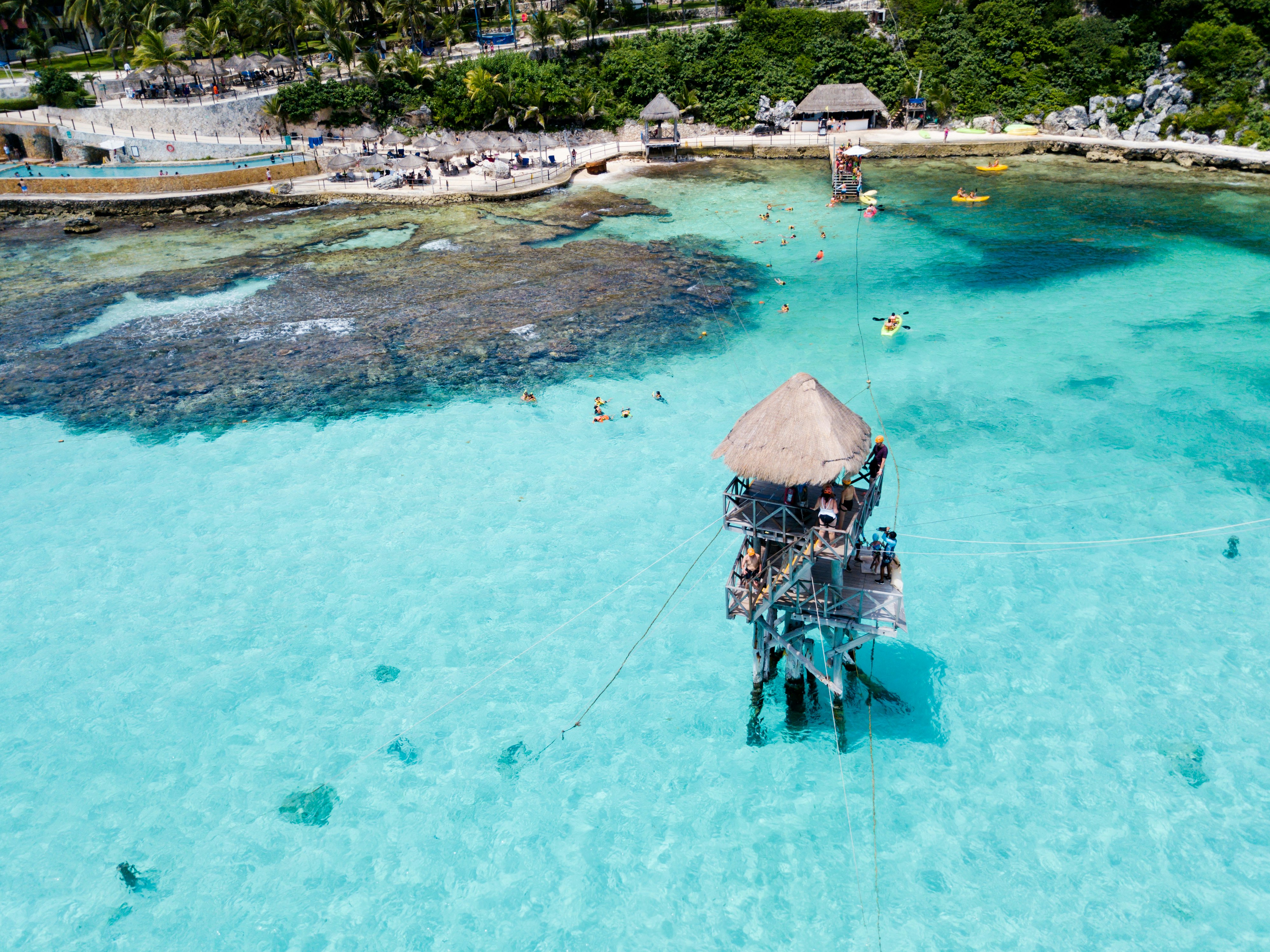 Aerial shot of a tall hut in the middle of the bright blue ocean in Isla Mujeres. There are people on kayaks and others walking on the paved street. 