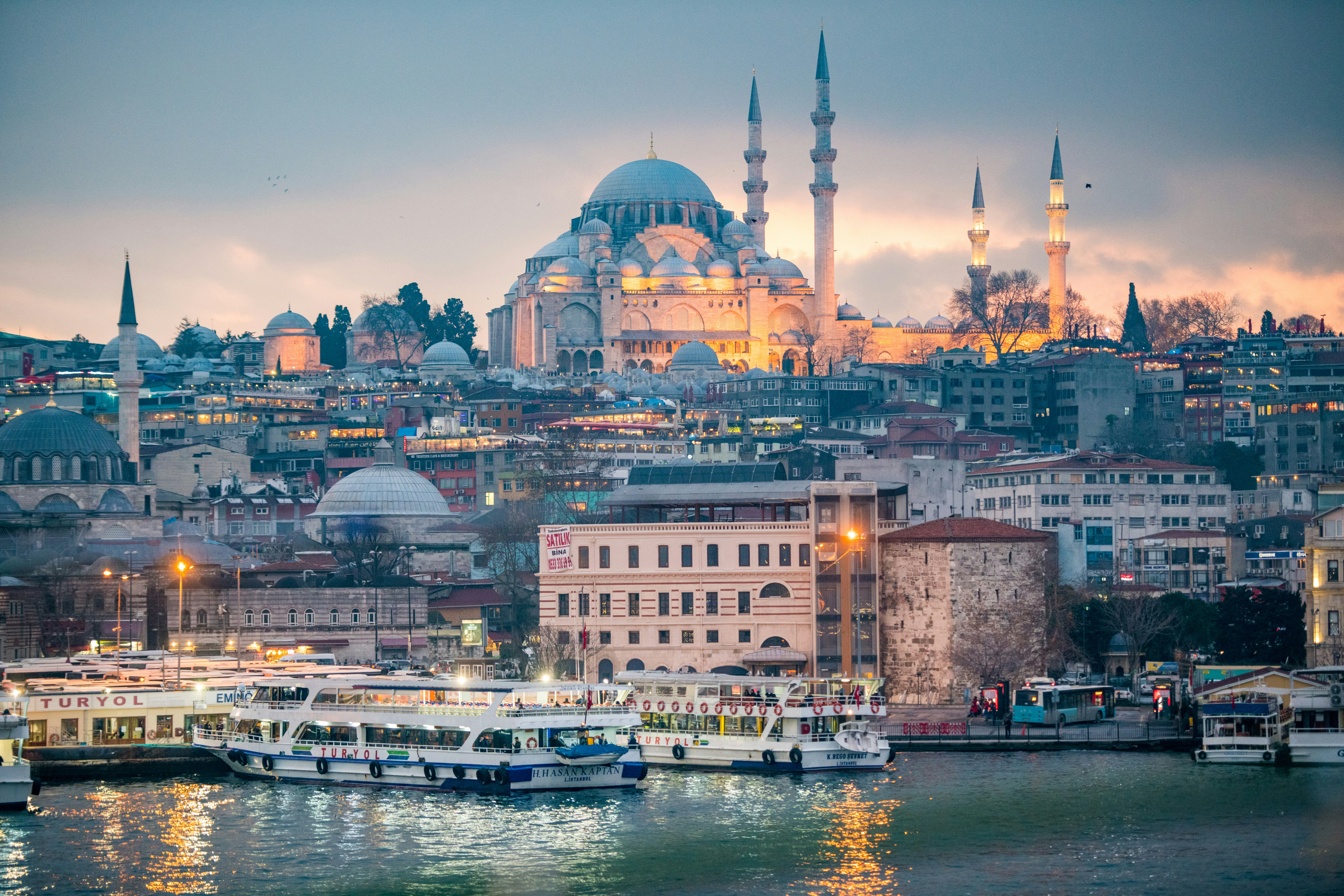 The glowing Süleymaniye Mosque sits on top of a hill overlooking the the city of Istanbul, Turkey, as street lights begin to illuminate the twilight skies. In front, a series of boats are visible.