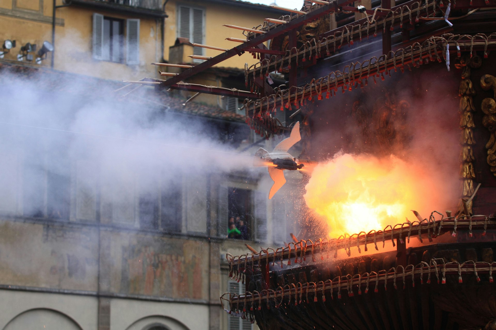 A mechanical bird is mid-flight, heading towards a flaming structure during Florence's Scoppio del Carro Easter celebration.