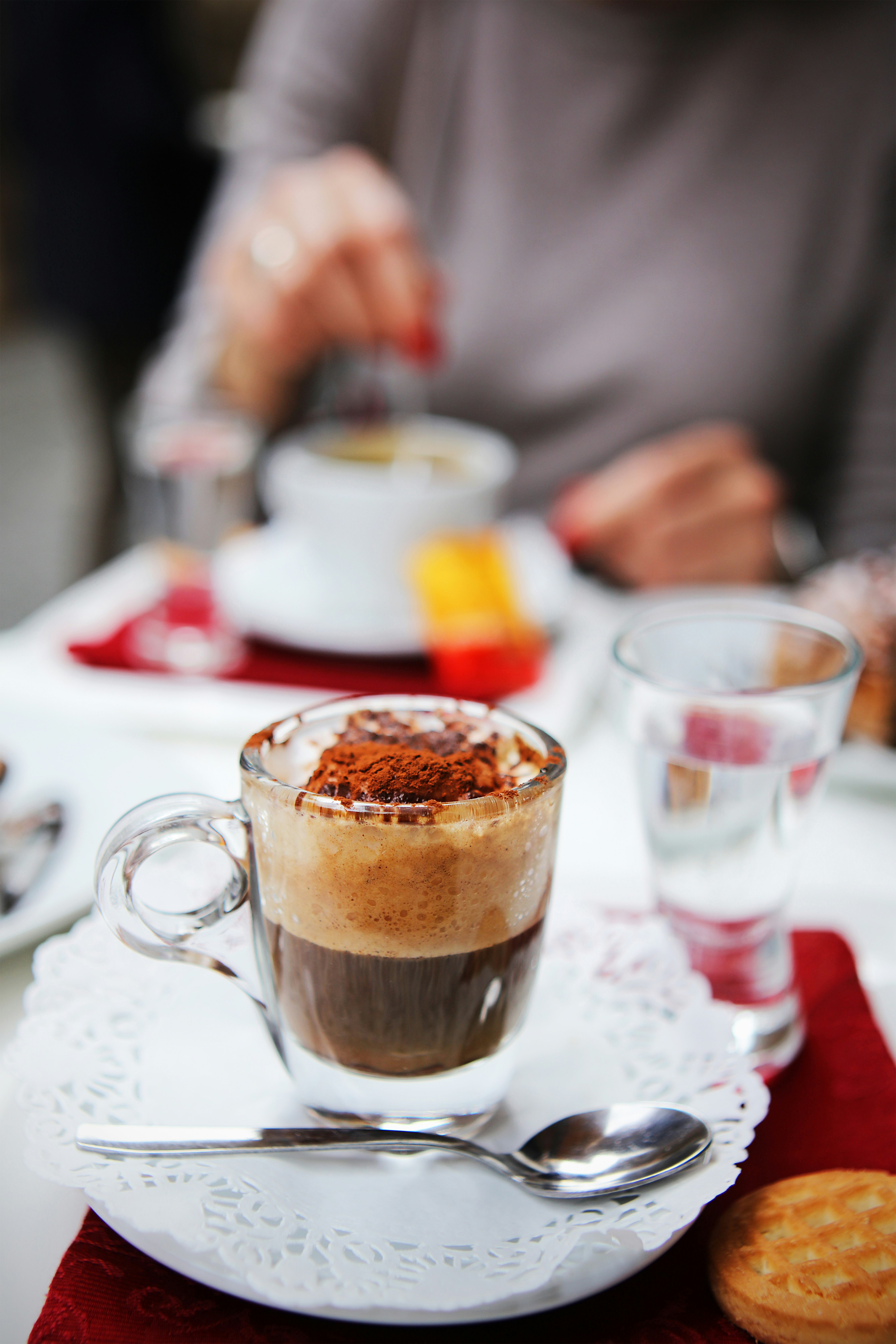 A close up shot of a cappuccino served in a class cup with a handle, on a white paper doily. There is a person wearing red nail polish stirring another cup of coffee out of focus in the background. 