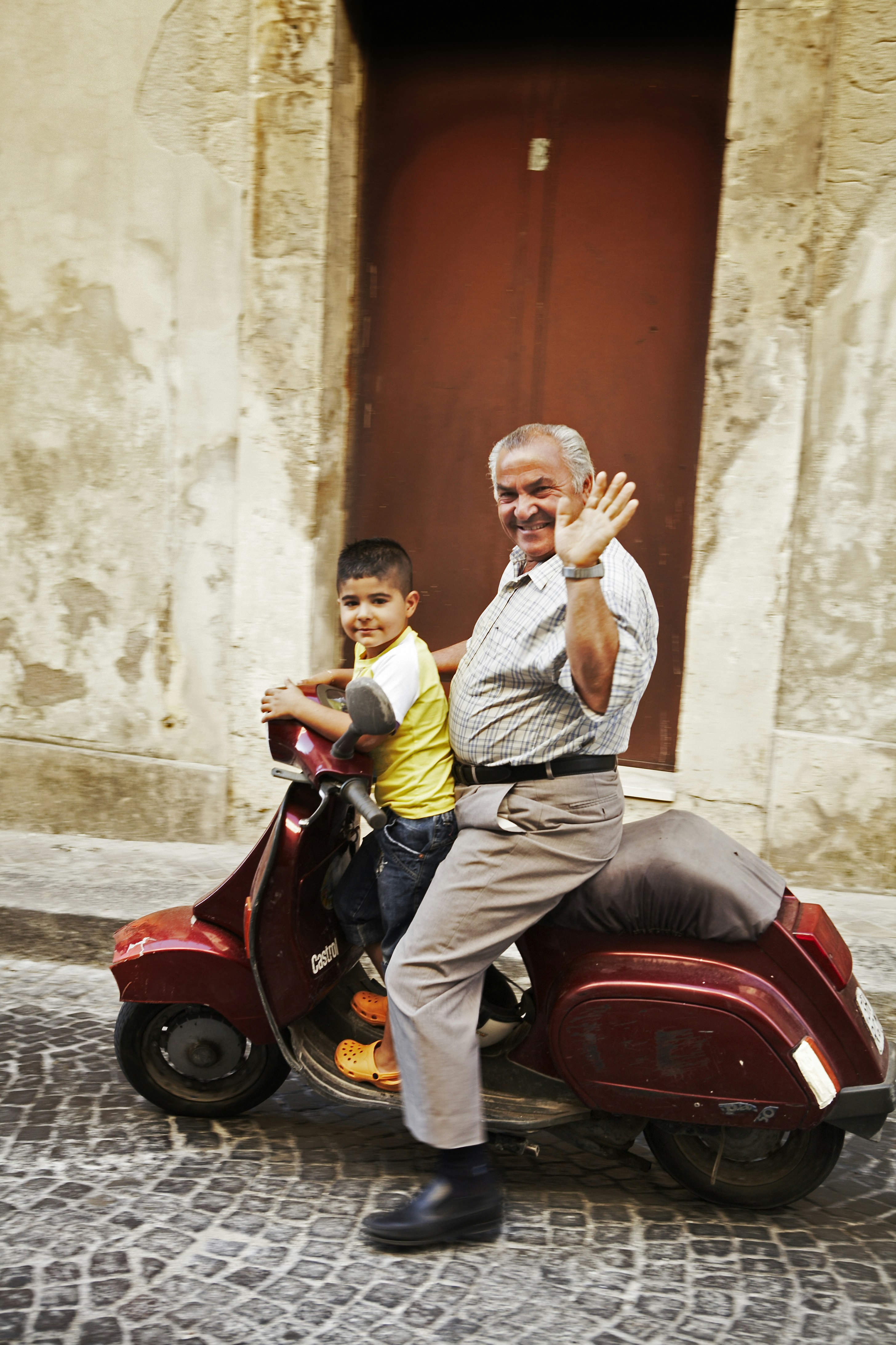 Man with child waving from Vespa motor scooter. The child is wearing orange crocs, shorts nd a yellow t-shirt. He is standing in front of an older man who sits on the scooter, waving at the camera, wearing a checked shirt and beige slacks. 