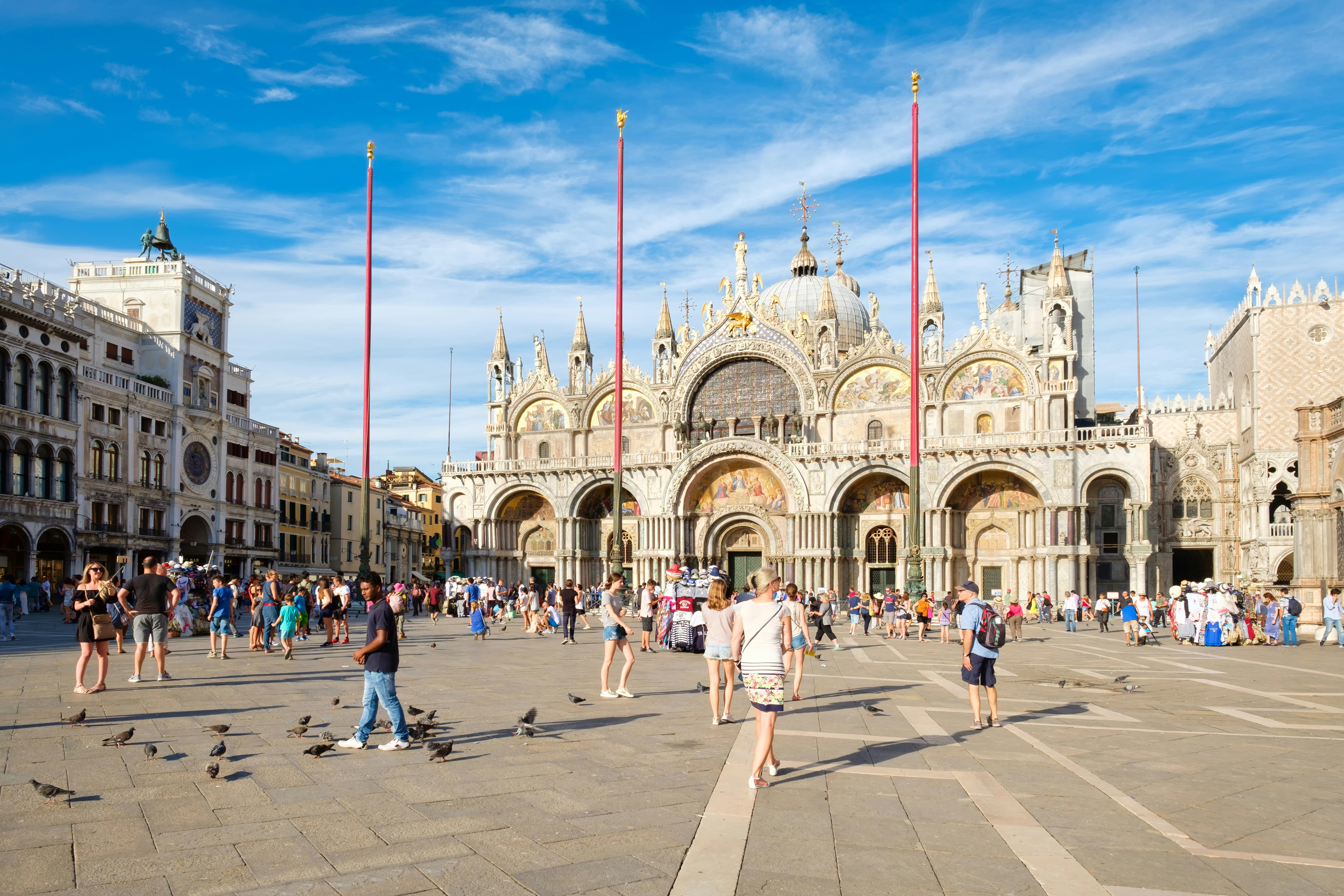 St Mark's Square in Venice on a sunny summer day. There are a few people and pigeons in the large tiled square and St Mark's Basilica is in the background. 
