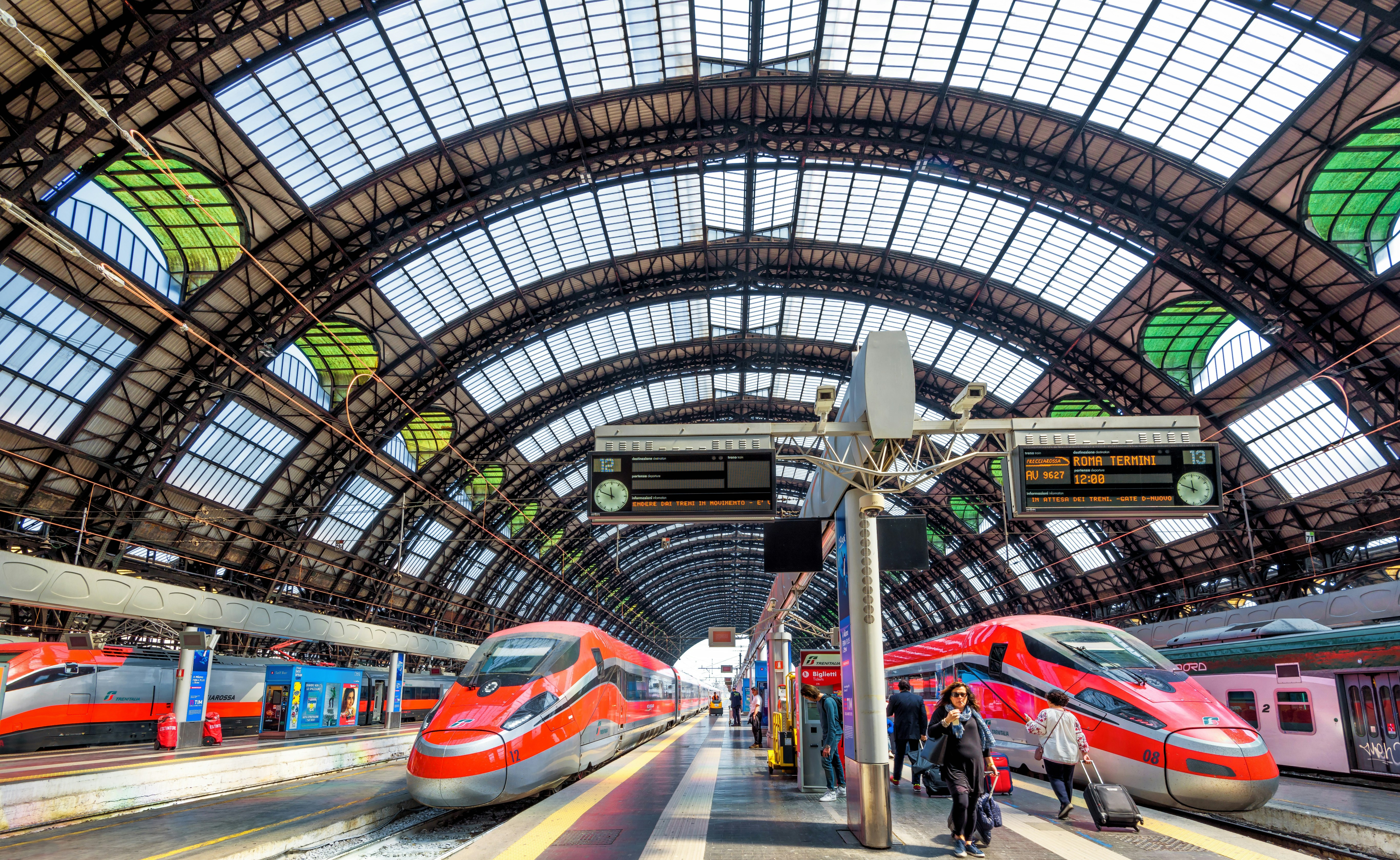 Modern high-speed trains at the railway Milan Central Station. The sleek, red and blue trains sit beneath a domed glass ceiling that has see-through panes with green accents. 