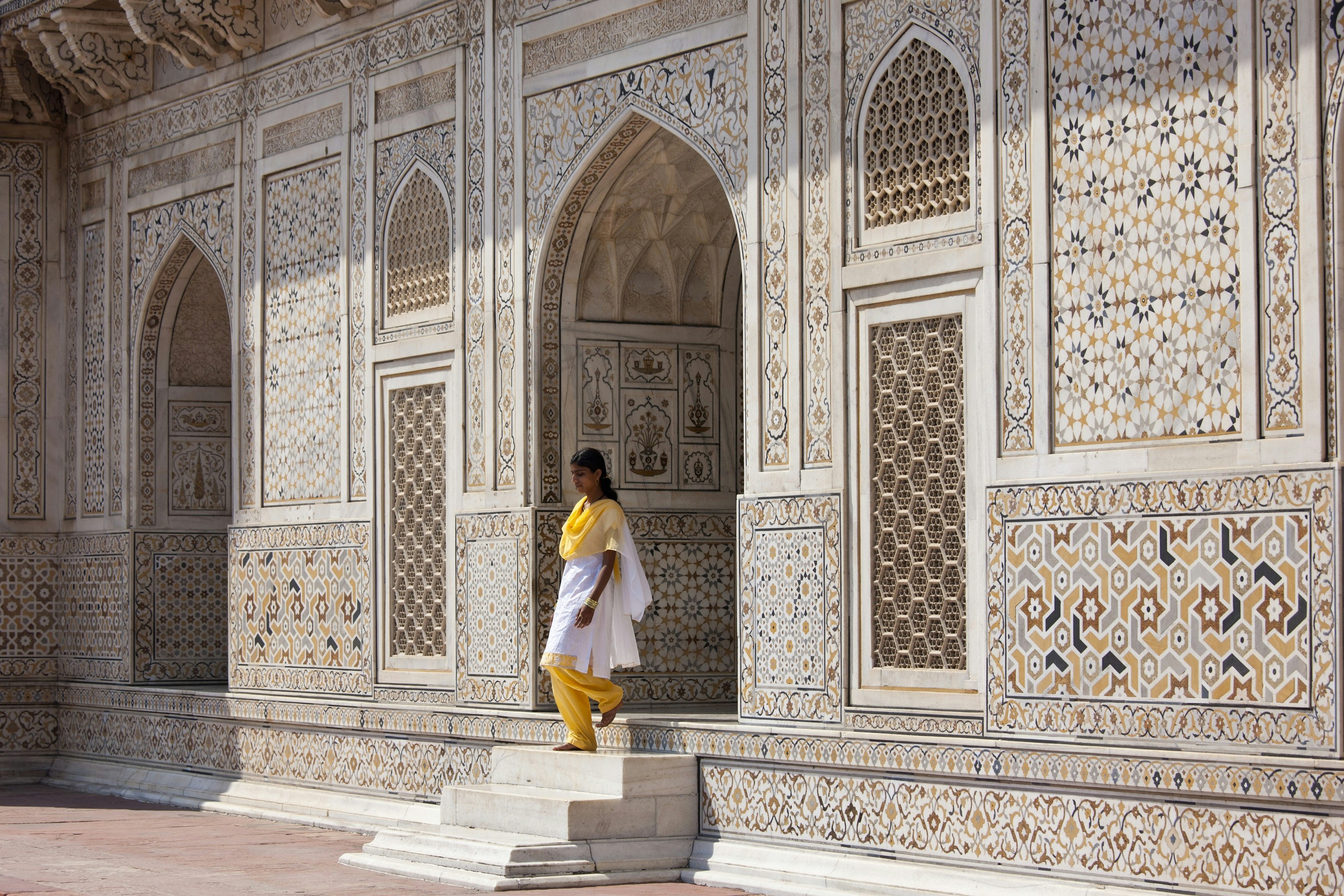 A woman in a yellow sari walks down the steps from the entrance of Itmad-ud-Daulah, a mausoleum in Agra. The white mausoleum is covered in colourful tiles.