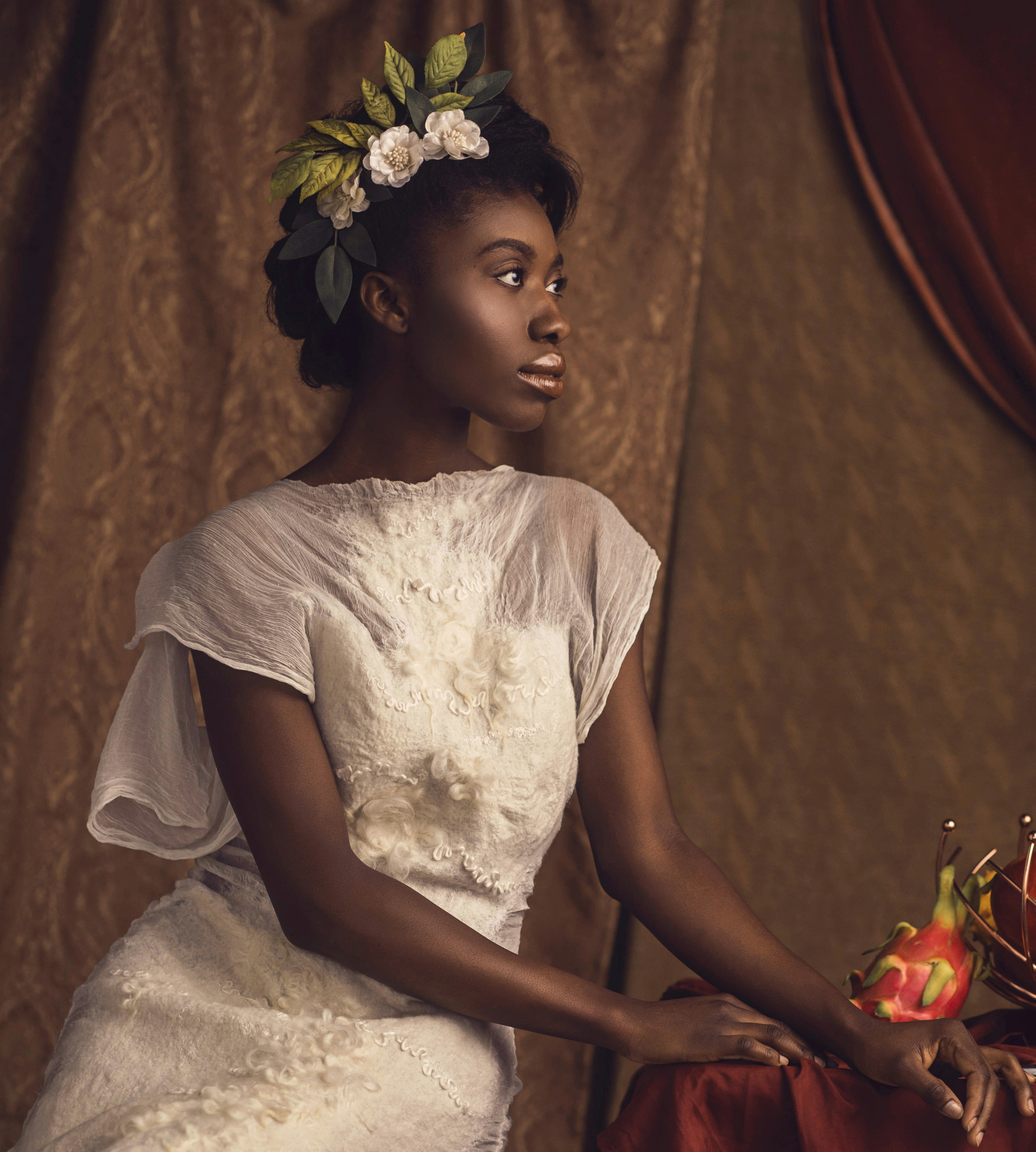 A fashion model with dark brown skin and a crown of white flowers and green leaves tucked into her updo sits partially in profile in one of Celeste Malvar-Stewart's white couture wedding gowns made from felted wool. The model is in front of a light brown damask backdrop and her hands rest on a table where a pink dragonfruit is displayed. 