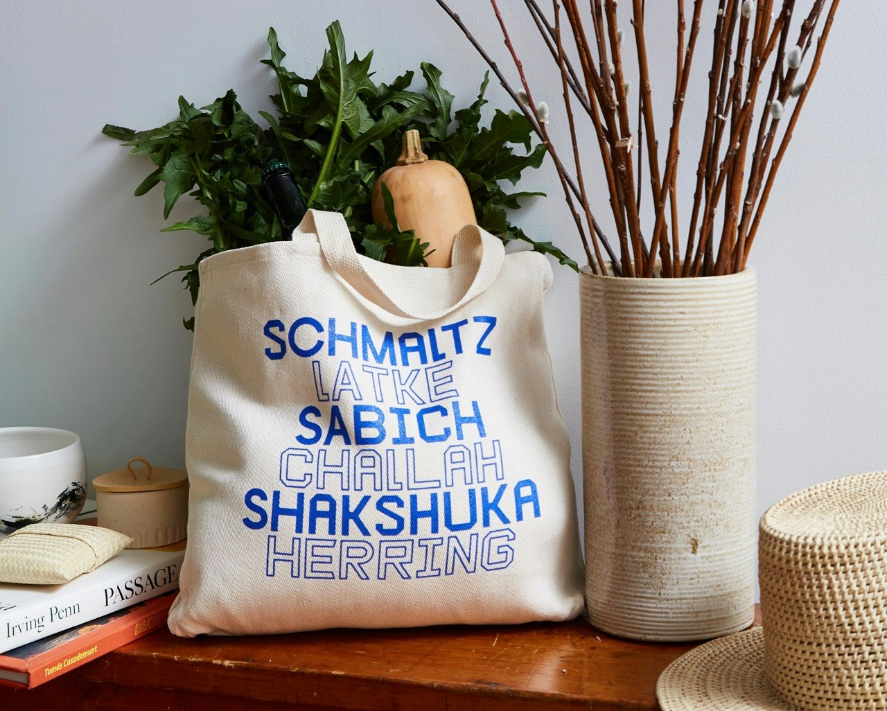 Canvas tote with names of Jewish foods in blue, filled with produce and sitting on a bench