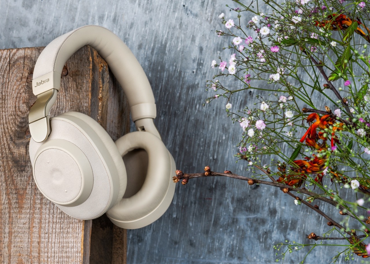 Jabra Elite 85h Gold Beige hanging from a piece of wood in front of a blue-gray background with flowers to the right