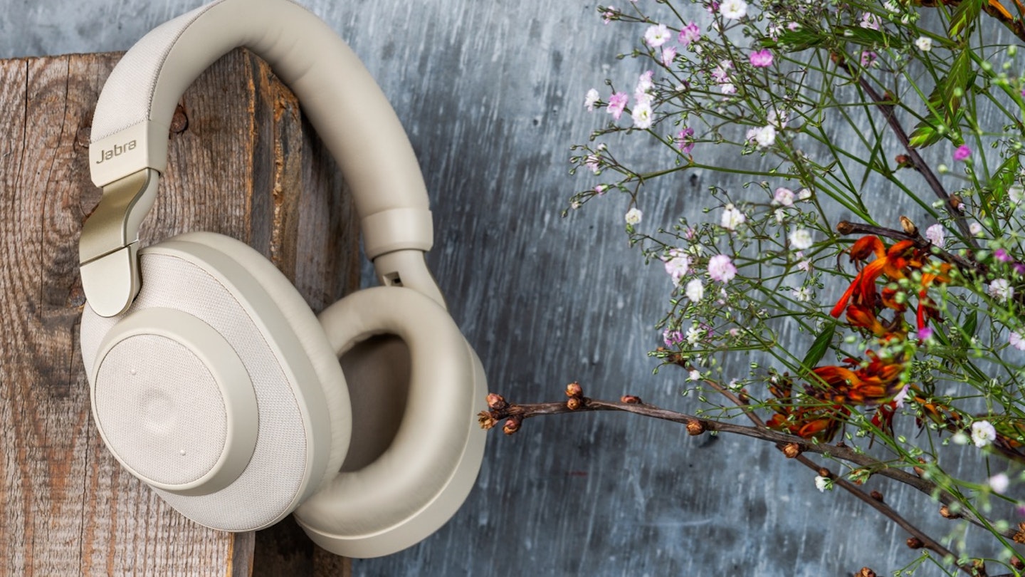 Jabra Elite 85h Gold Beige hanging from a piece of wood in front of a blue-gray background with flowers to the right