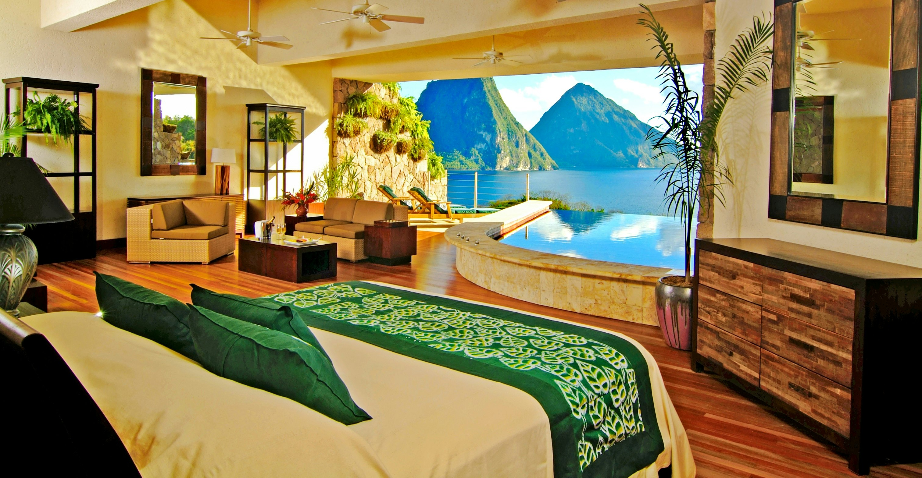 The foreground is filled with a bed with white sheets and emerald green pillows, as well as a runner with a leaf motif at the foot of the bed. It faces an expansive opening the width of the room overlooking mountains rising out of the ocean. In one corner of the room is a living room arrangement with a coffee table, love seat, and easy chair. Directly in front of the bed is a curved in-room infinity pool also overlooking the sea.