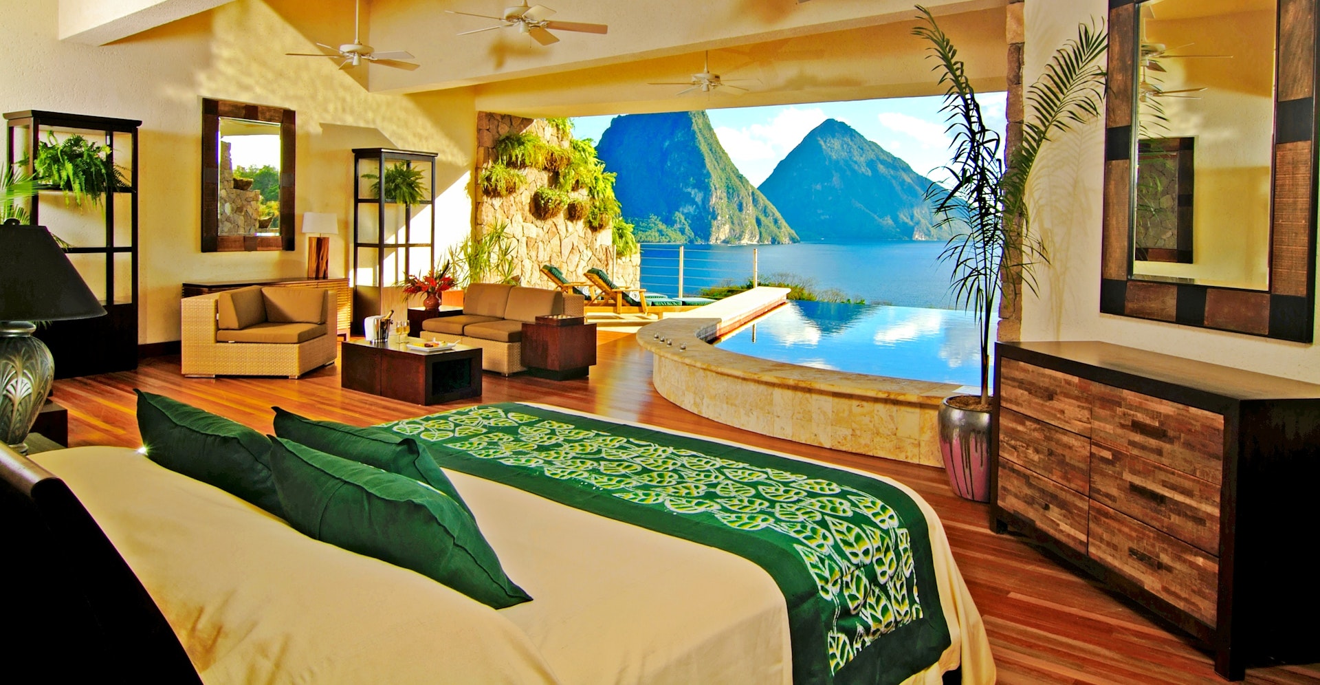 The foreground is filled with a bed with white sheets and emerald green pillows, as well as a runner with a leaf motif at the foot of the bed. It faces an expansive opening the width of the room overlooking mountains rising out of the ocean. In one corner of the room is a living room arrangement with a coffee table, love seat, and easy chair. Directly in front of the bed is a curved in-room infinity pool also overlooking the sea.