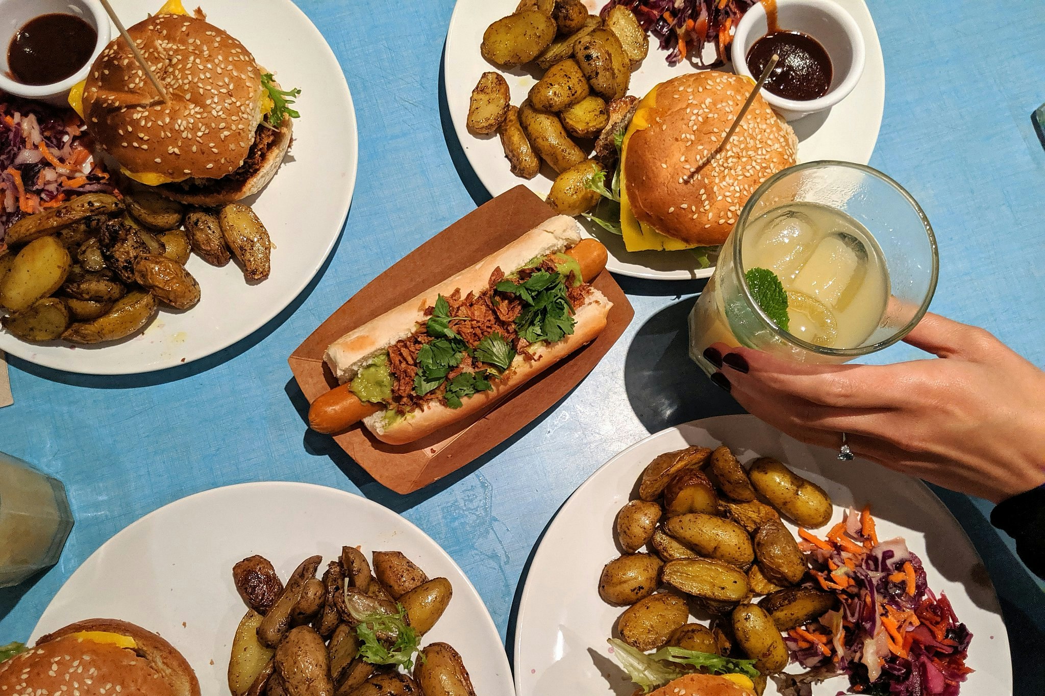 Plates of vegan burgers with potatoes and slaw, plus a vegan hot dog, on a blue tabletop.