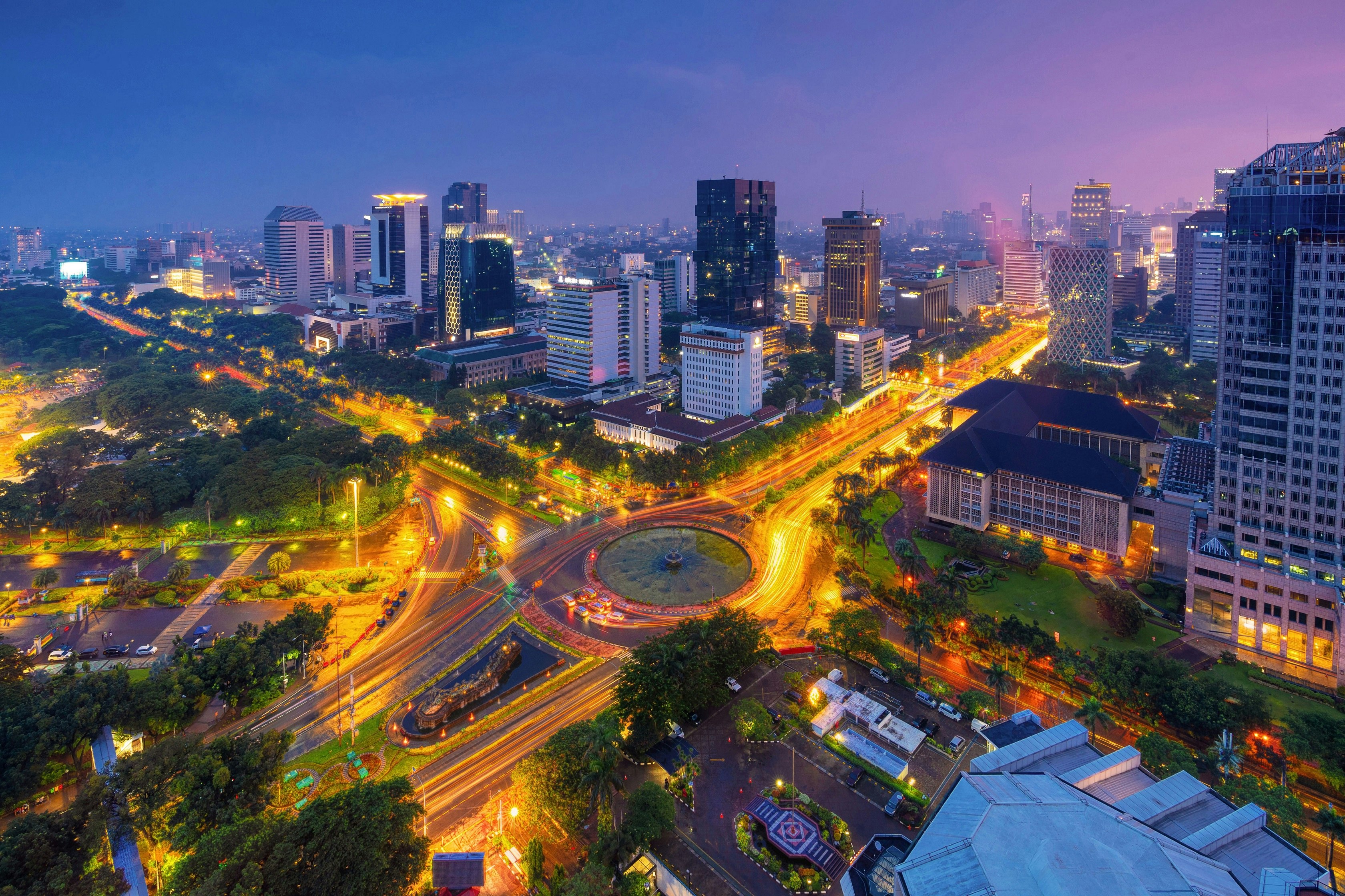An aerial view of the city of Jakarta taken at night. The windows of skyscrapers are illuminated in the darkness while light trails show the movement of traffic around the busy streets.