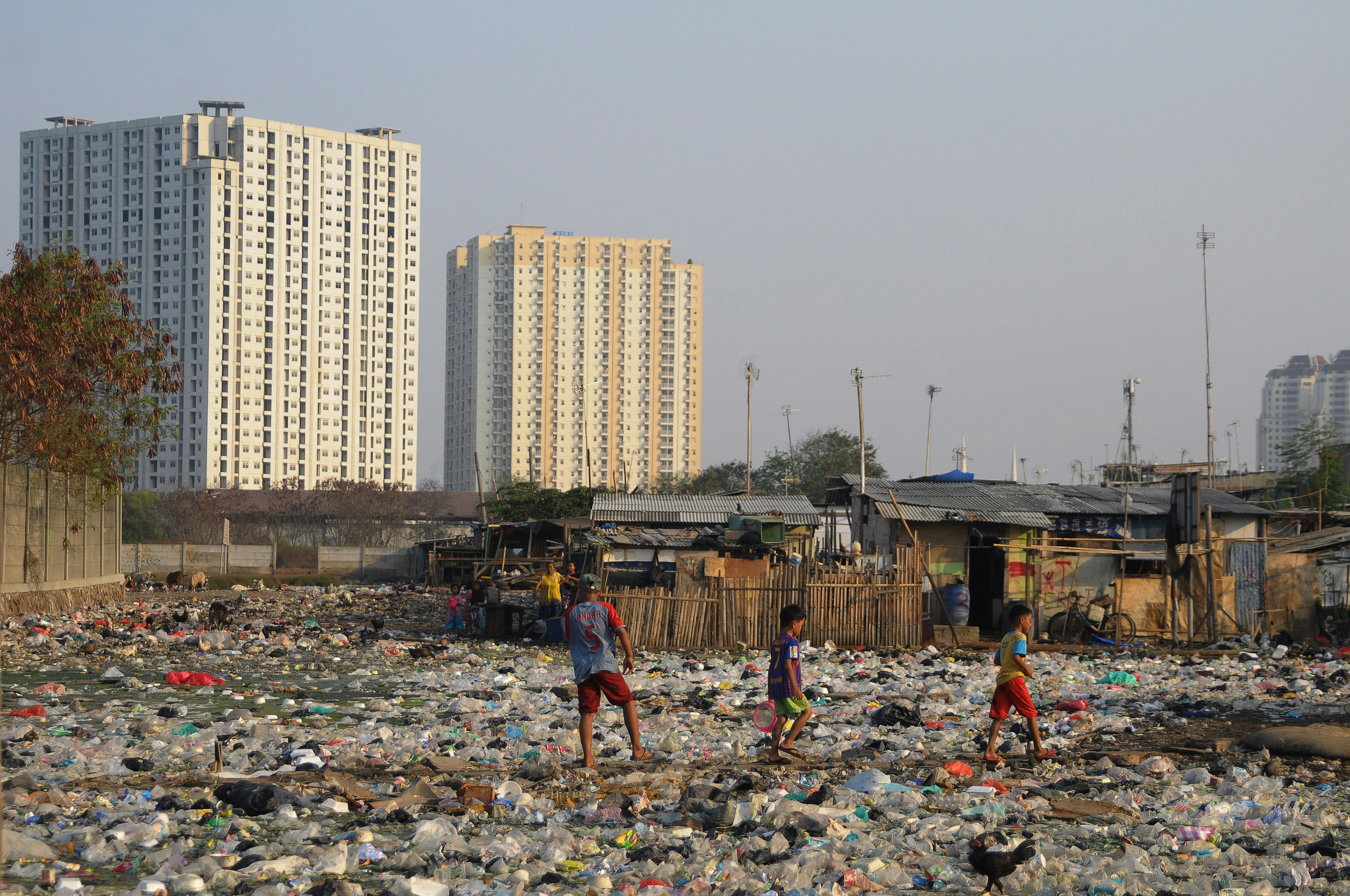 Plastic waste and pollution is spread across a large open space on the poorer outskirts of Jakarta. In the background two high-rise buildings are visible.