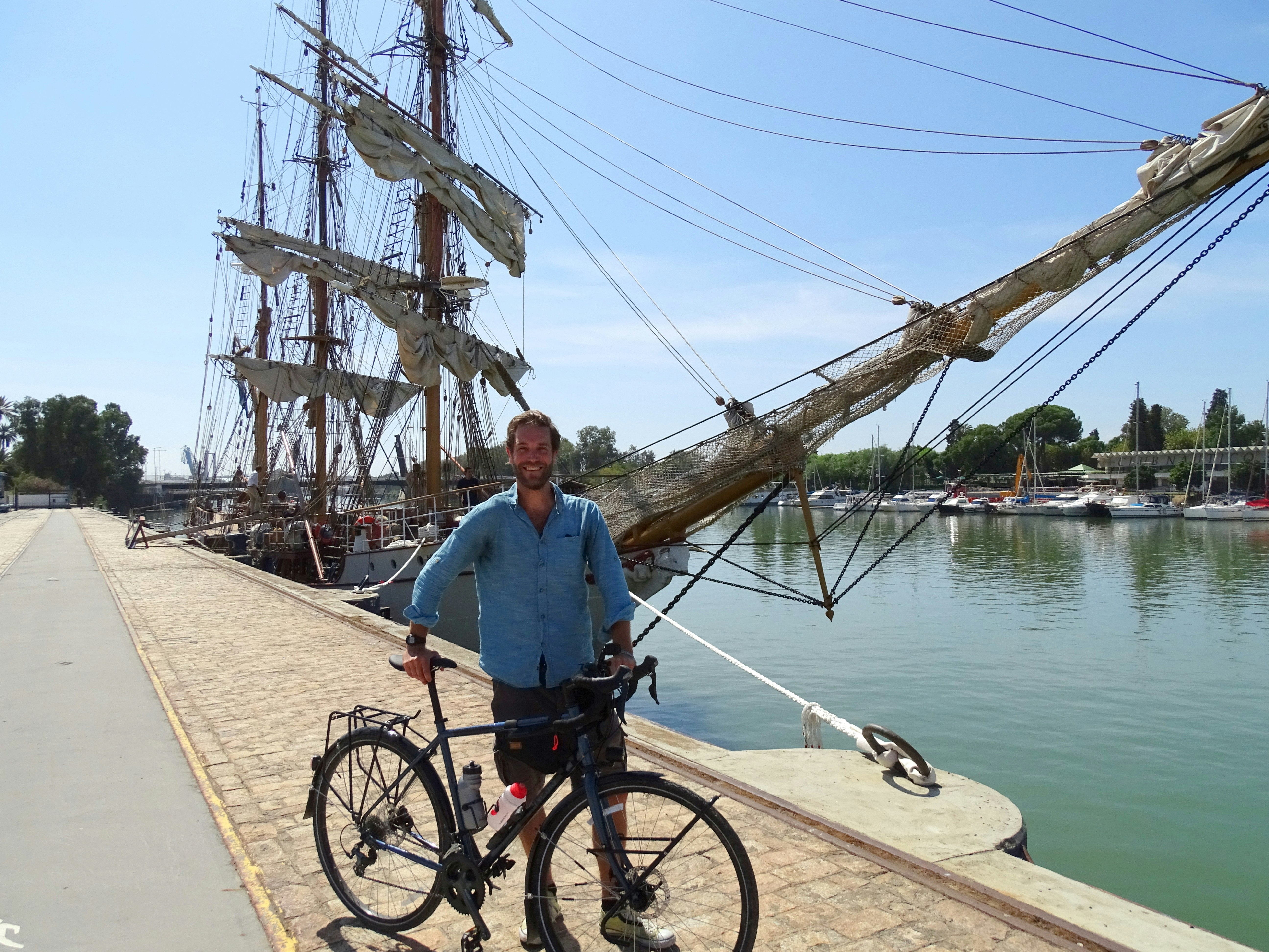 James stands with a bike on the water's edge next to a tall sailing ship. 