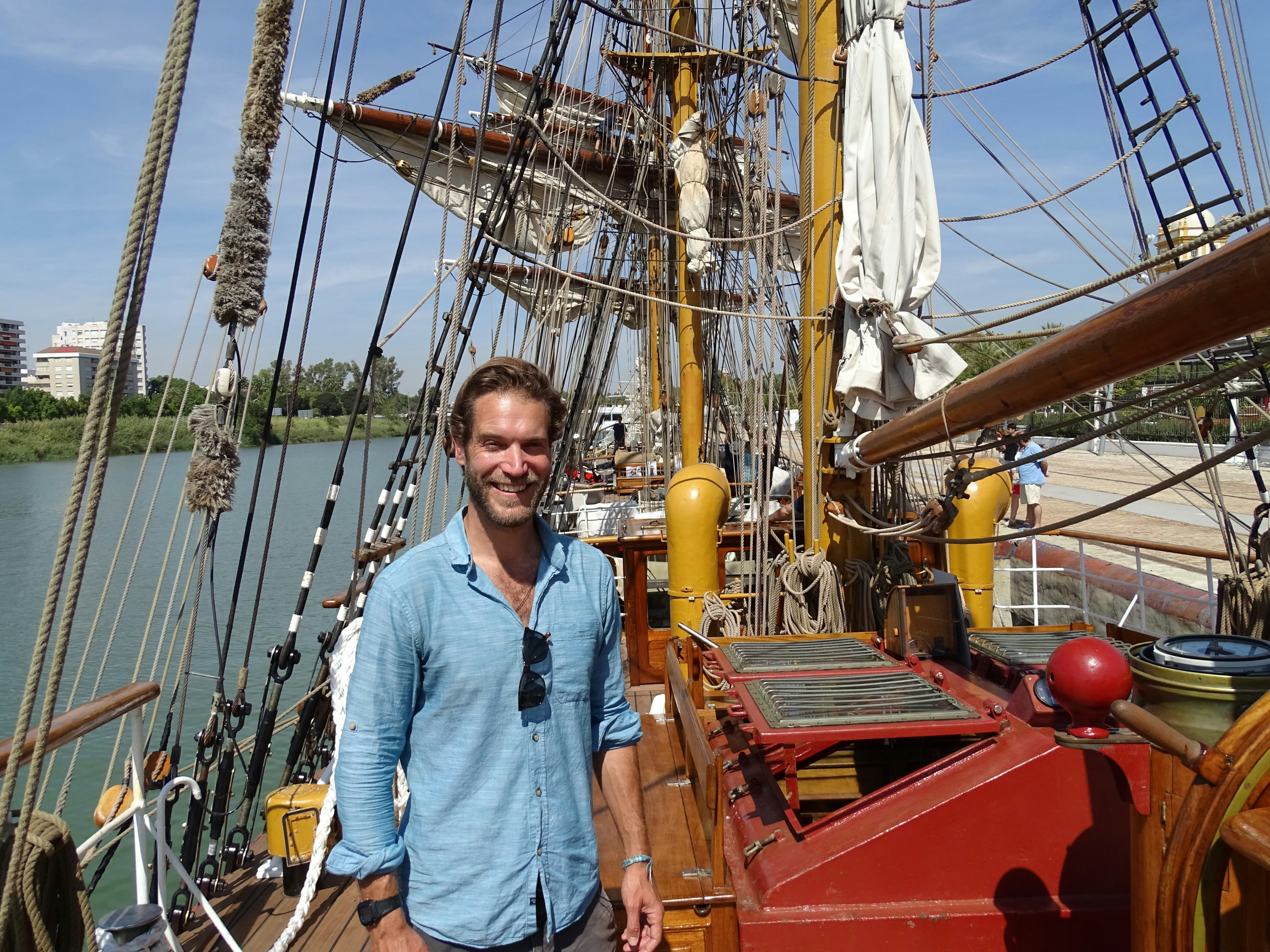 James, wearing a blue shirt, stands on board the tall ship. 