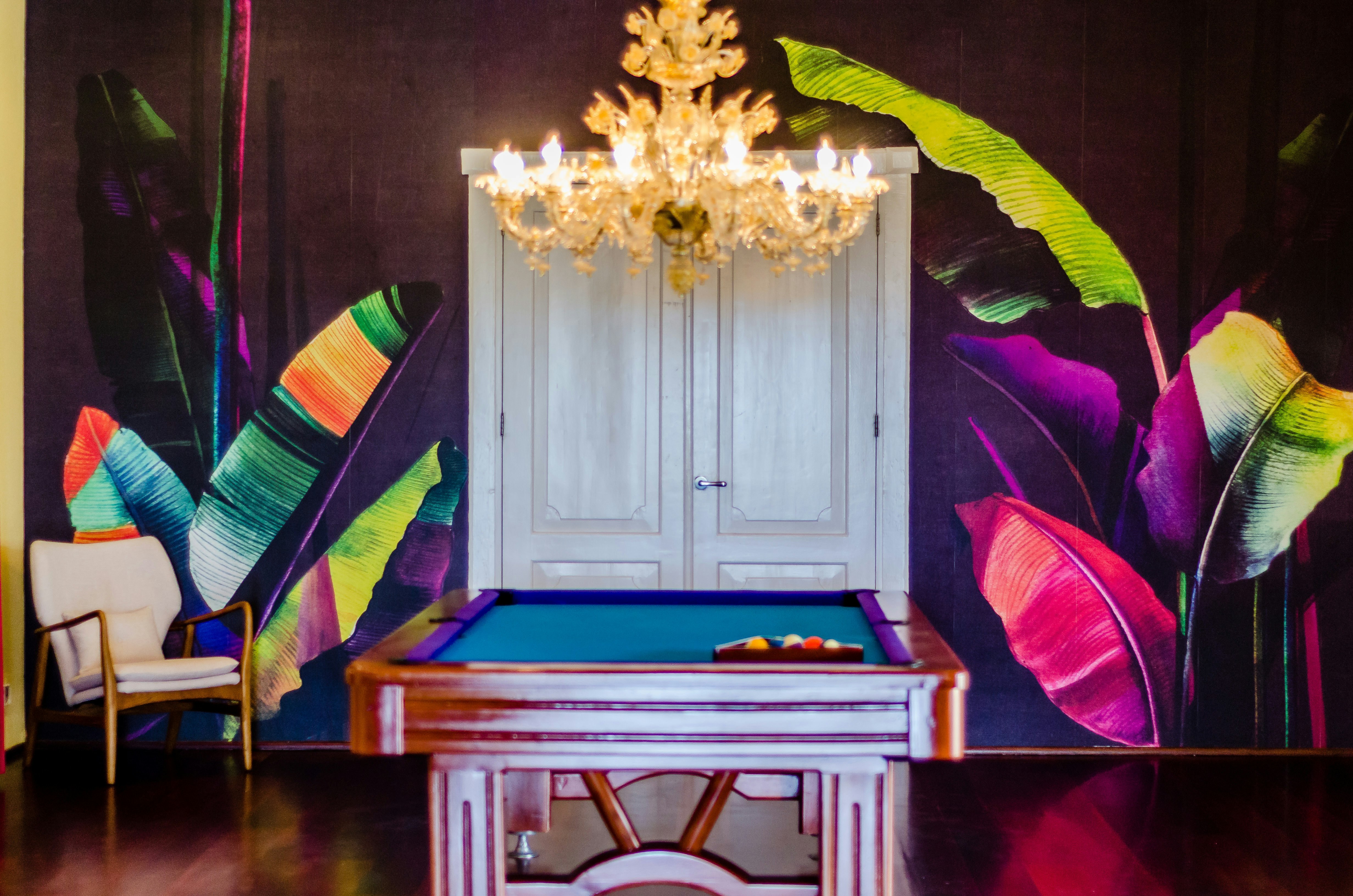 A pool table sits under a crystal chandelier in front of a deep purple wall decorated with a colorful illustration of tropical plants. A white midcentury modern chair sits in the corner and white French doors sit at the far end of the pool table