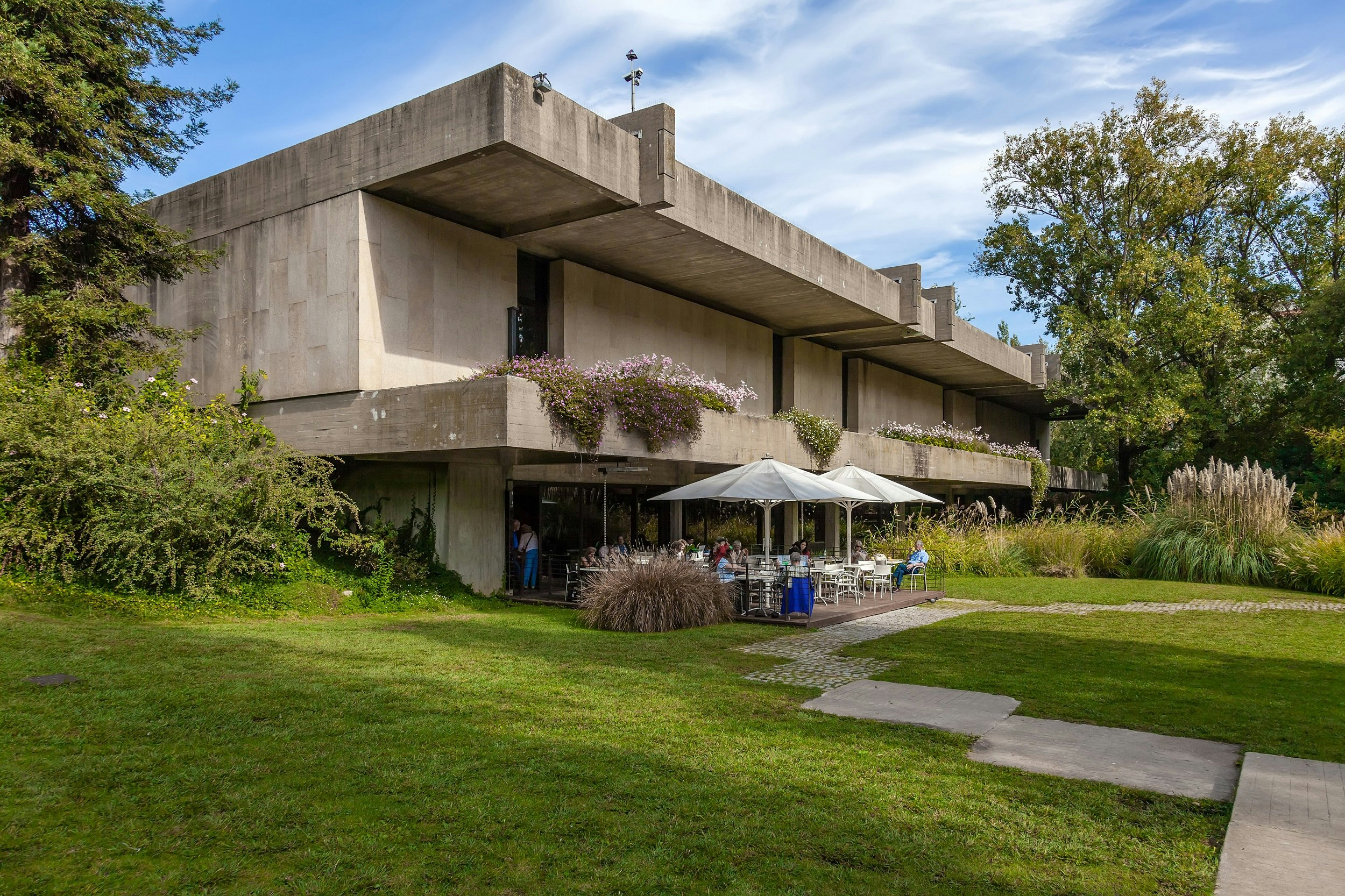 A manicured lawn and large trees surround the brutalist concrete structure of the Calouste Gulbenkian foundation; on the ground level is a cafe with table seating under large umbrellas on a patio.