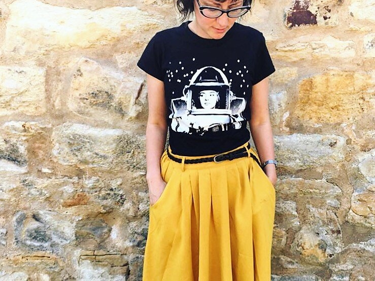 Jenny Elliott standing against a sandstone wall in a black t-shirt and yellow skirt