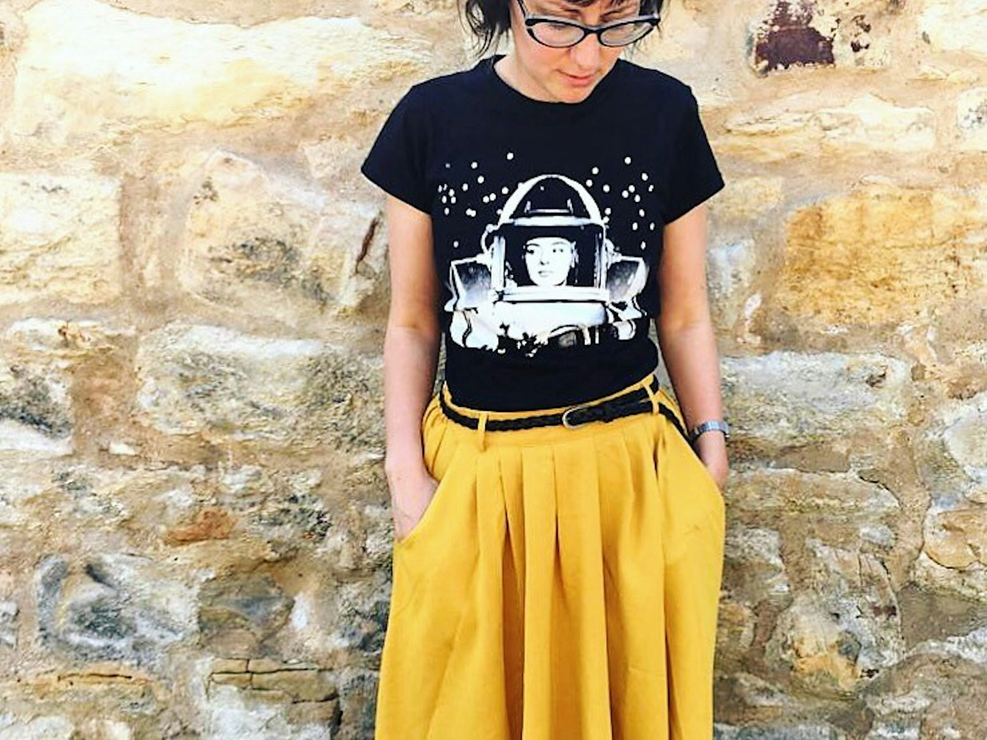 Jenny Elliott standing against a sandstone wall in a black t-shirt and yellow skirt