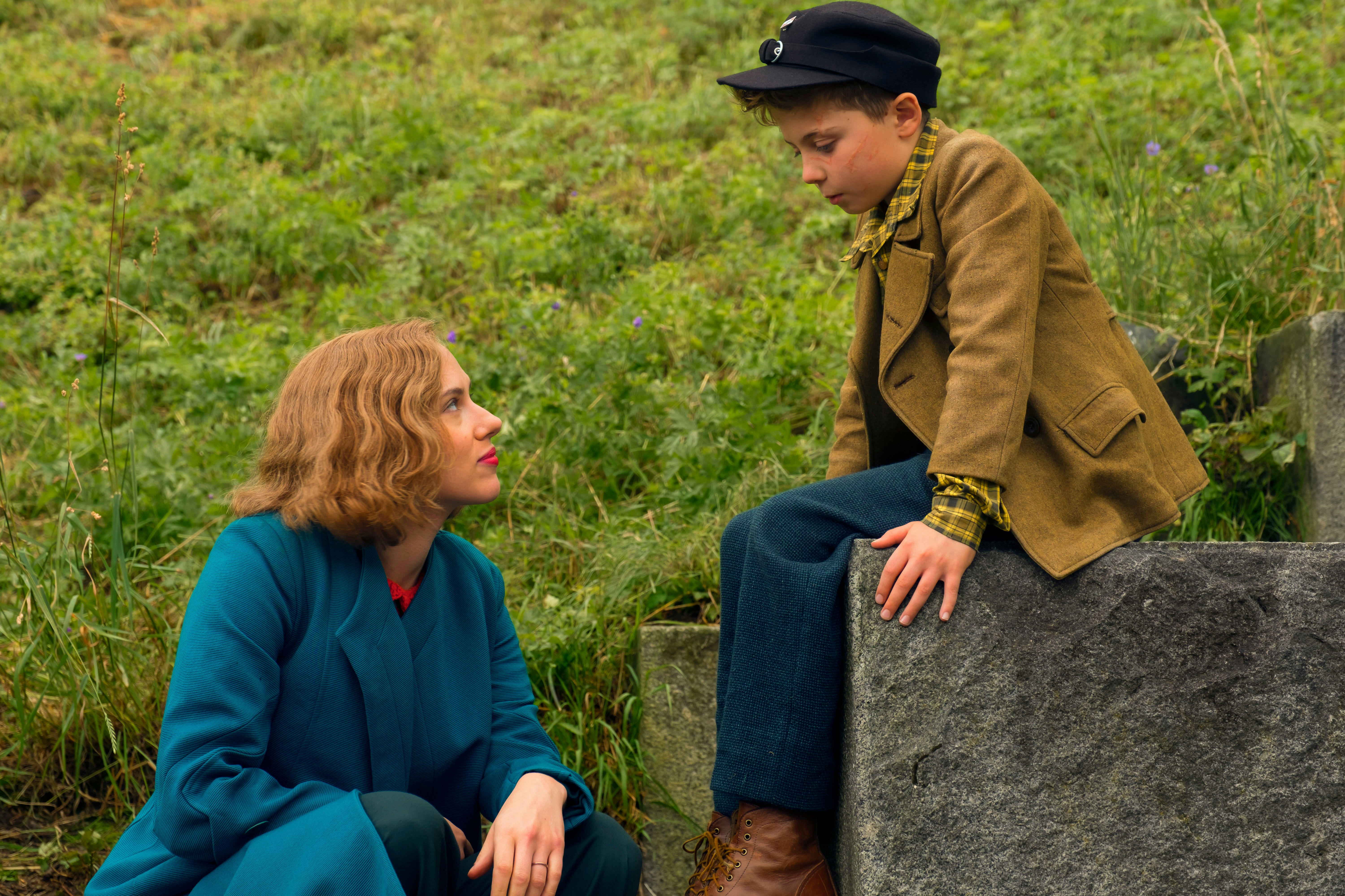 In a still from the movie 'Jojo Rabbit, actor Scarlett Johansson crouches down to talk to  Roman Griffin Davis, who is sitting on a large rock.