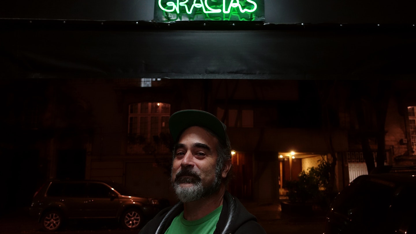 A man wearing a dark baseball hat, green t-shirt and black sweatshirt stands under a neon sign that says "Gracias".