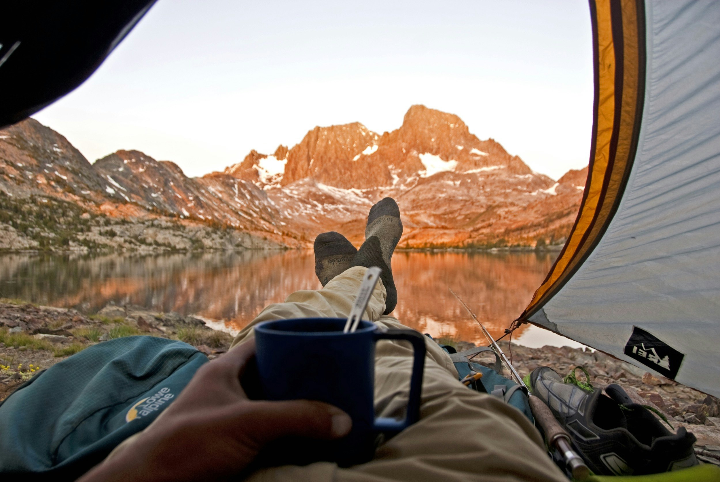 Looking out the entryway of a tent with the mountains of the Sierra Nevada in the distance, a man's feet and a cup of soup is balanced on his stomach.