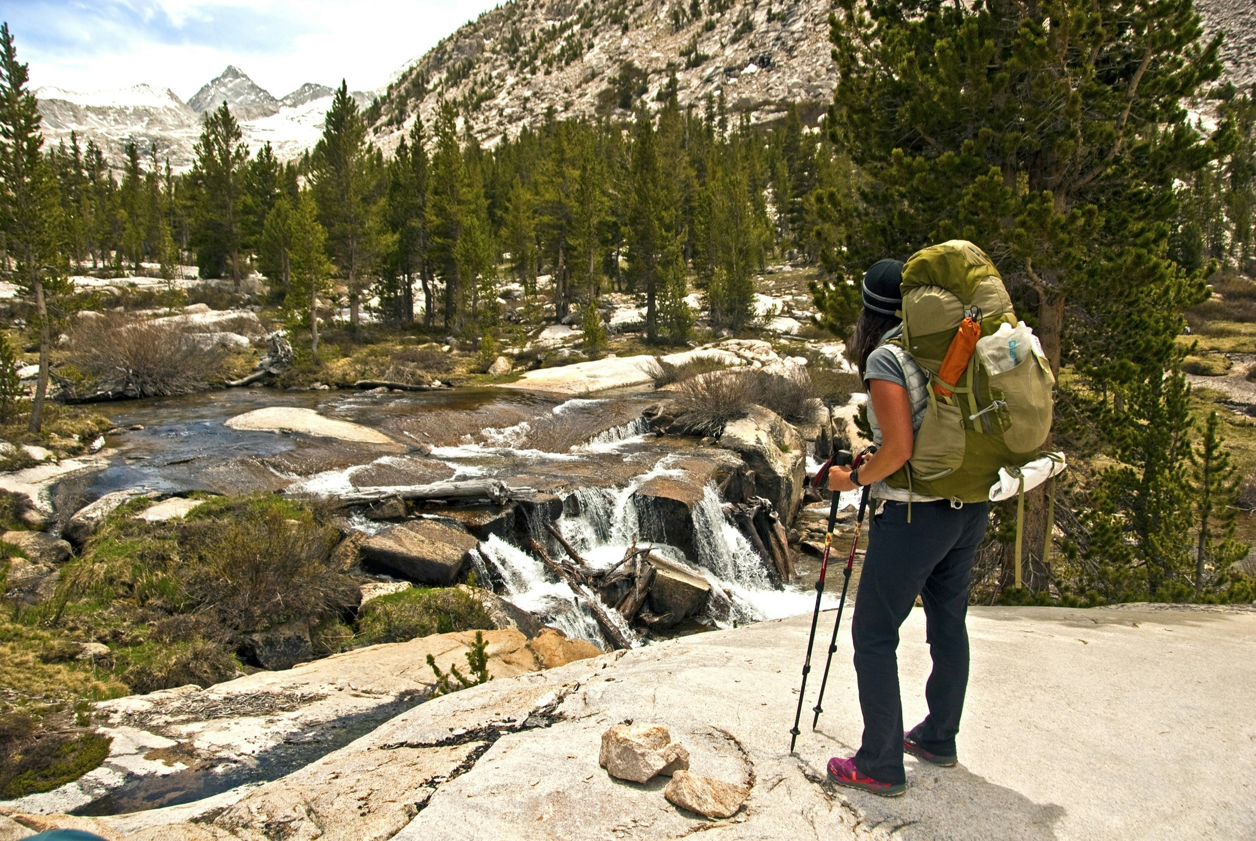 A woman stands on a large boulder as she looks at a tumbling creek at the base of a mountain on the John Muir Trail in California