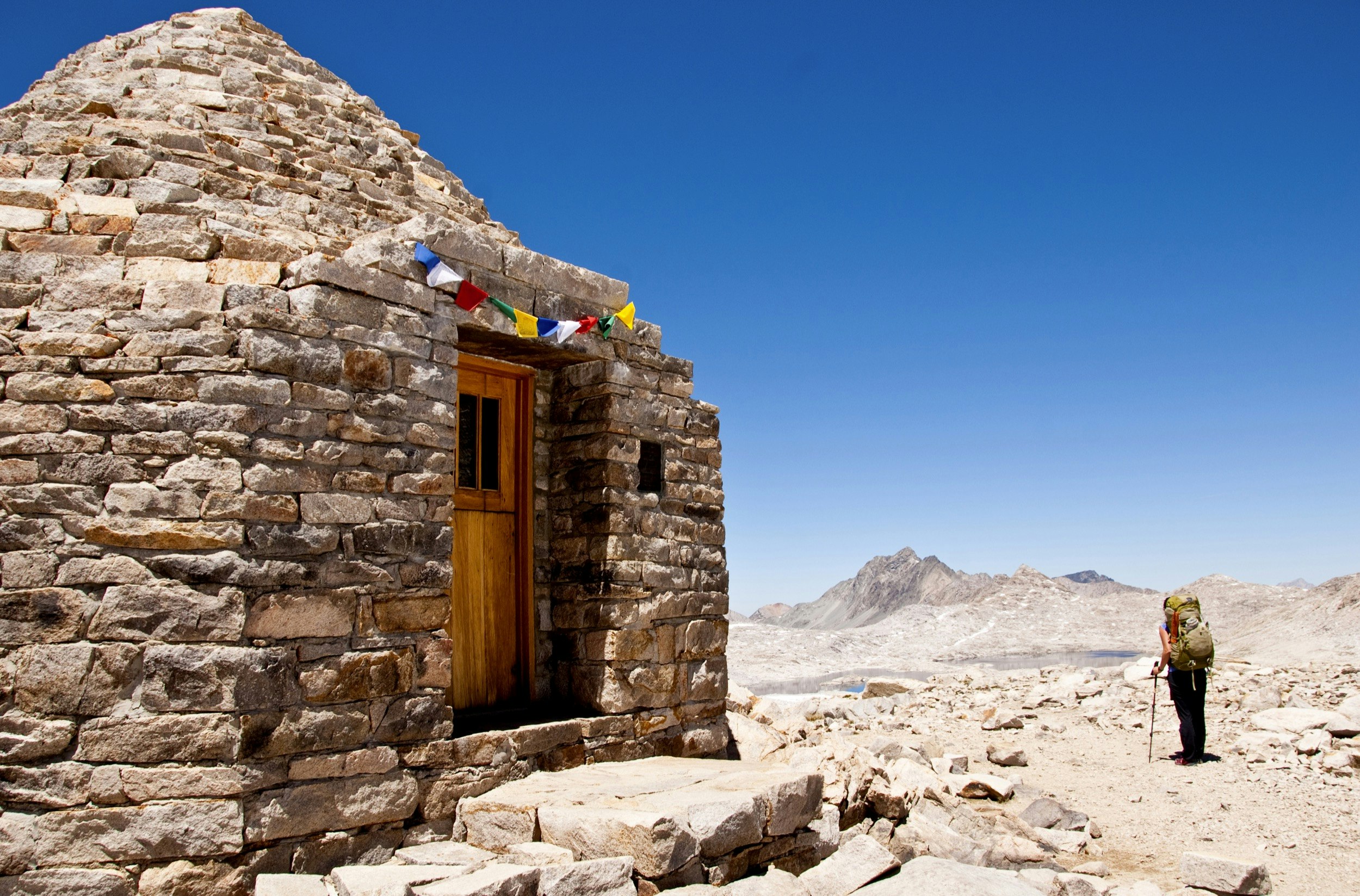 A woman stands next to small rustic stone shelter with colored flags over the door on the John Muir Trail in the Sierra Nevada mountain range