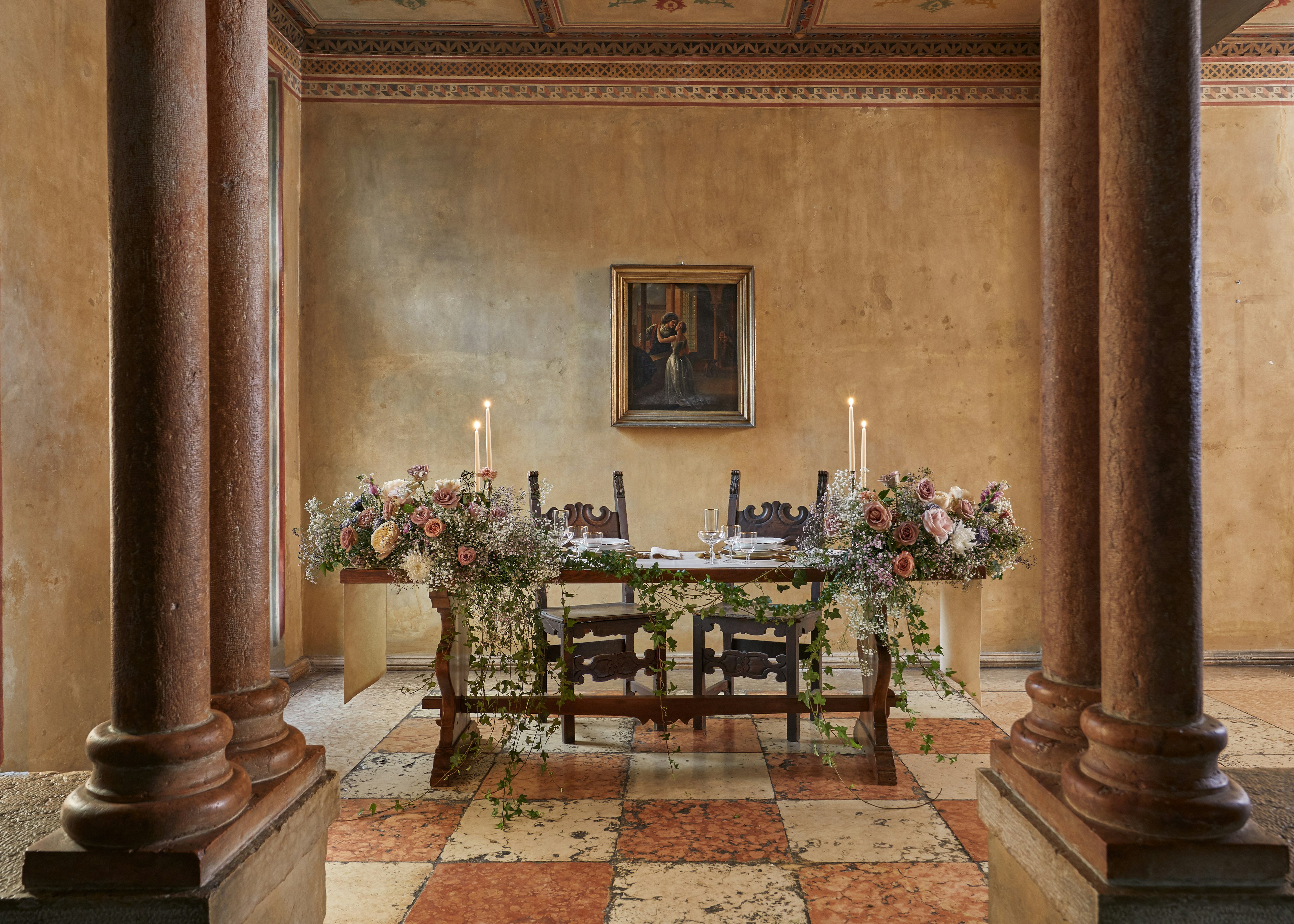 A dining table set with candles and and flowers, placed between columns on a checkerboard-tiled floor