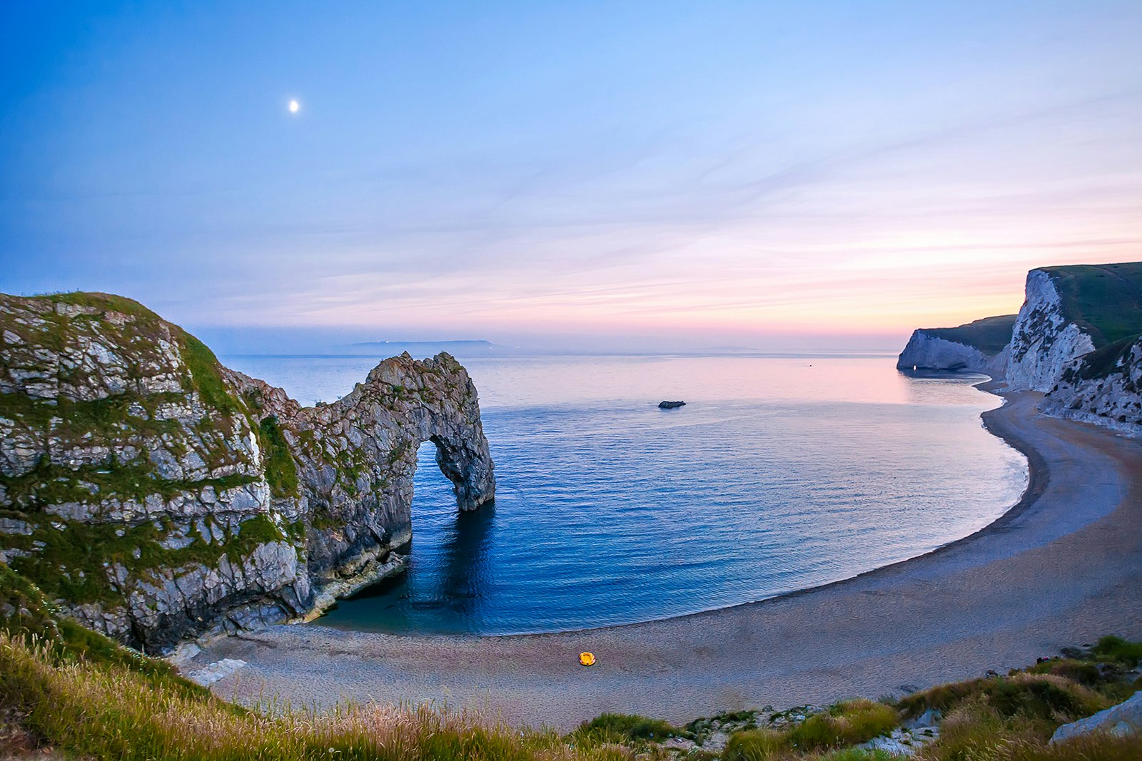A rock arch stretches into a small bay at twilight. A small yellow boat sits on the shore. Jurassic Coast, England.