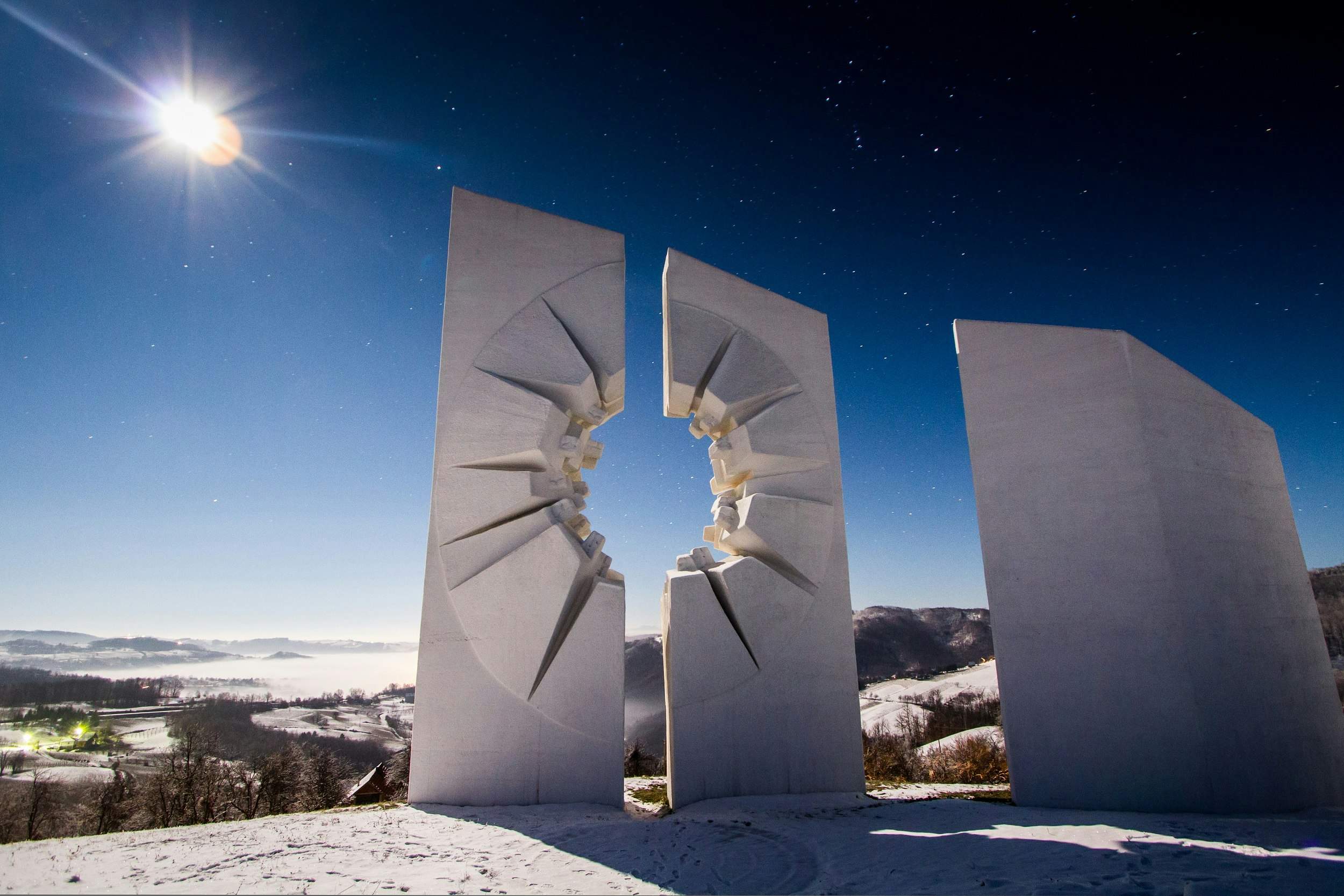 A large concrete monument with a hole in the centre of it, representing a bullet hole. Snowy fields can be seen in the background and the sun is beaming down