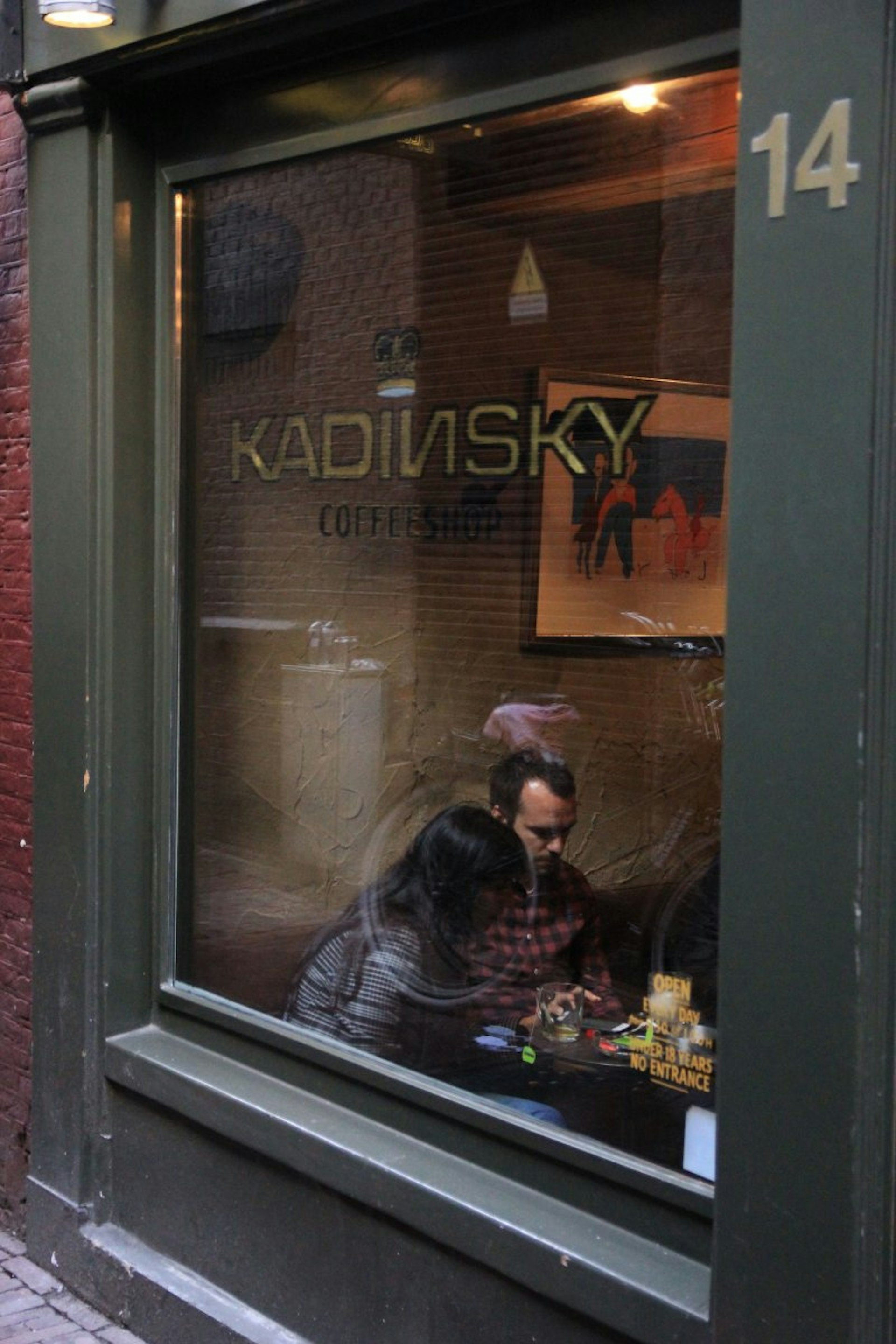 The exterior of Kadinsky Cafe Zoutsteeg which is painted black. Through the large window we can see a couple of people sitting at a table with drinks and art on the wall. 