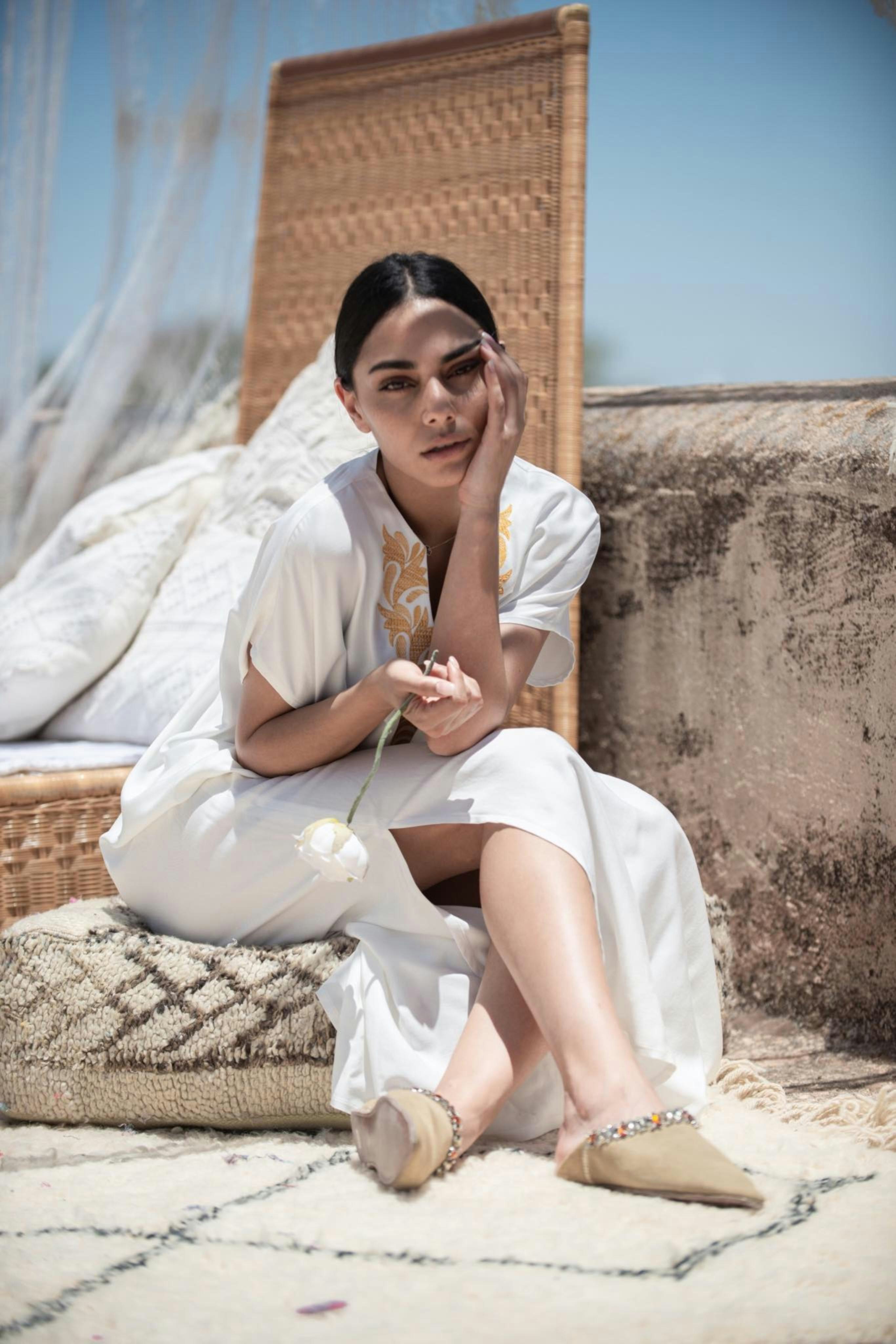 A model sits on a cushion on a rooftop wearing flowing, white-and-gold fabrics