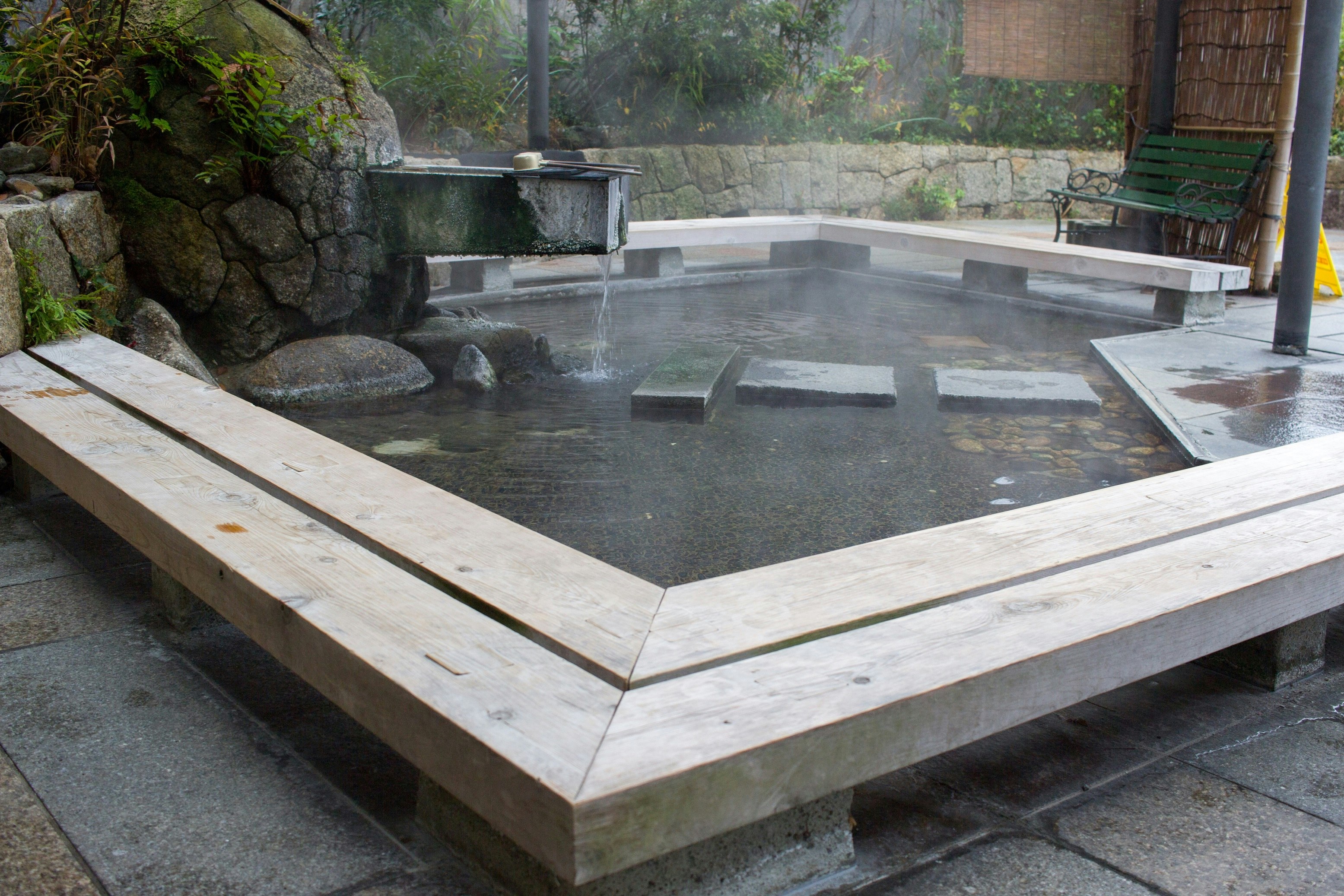 An onsen in Kaga. The hot pool is a hexagonal shape, with benches surrounding it on all sides. The water trickles in from a pipe above the pool.