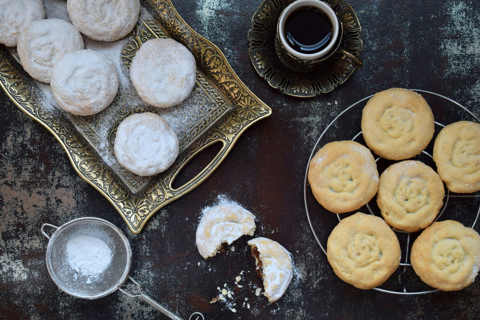 Sesame shortbread kahk cookes with date stuffing from Egypt. Some of the cookies are on a decorative gold tray sprinkled with icing sugar, one is broken in half to show the filling and the rest are on a cooling rack next to a small cup of tea.