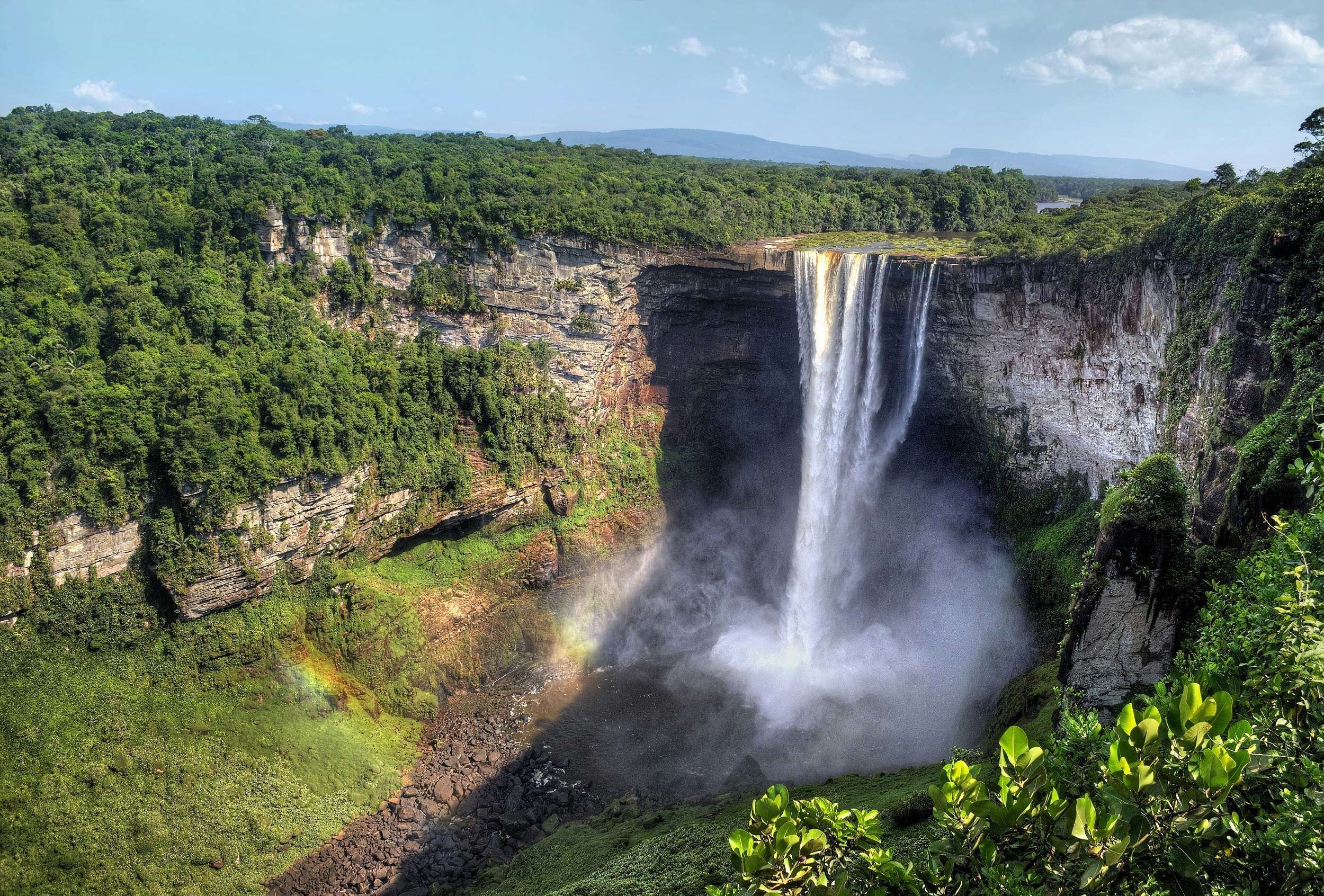 A large river plummets off a sheer white cliff into a mist filled canyon; the surrounding area is covered in lush rainforest.