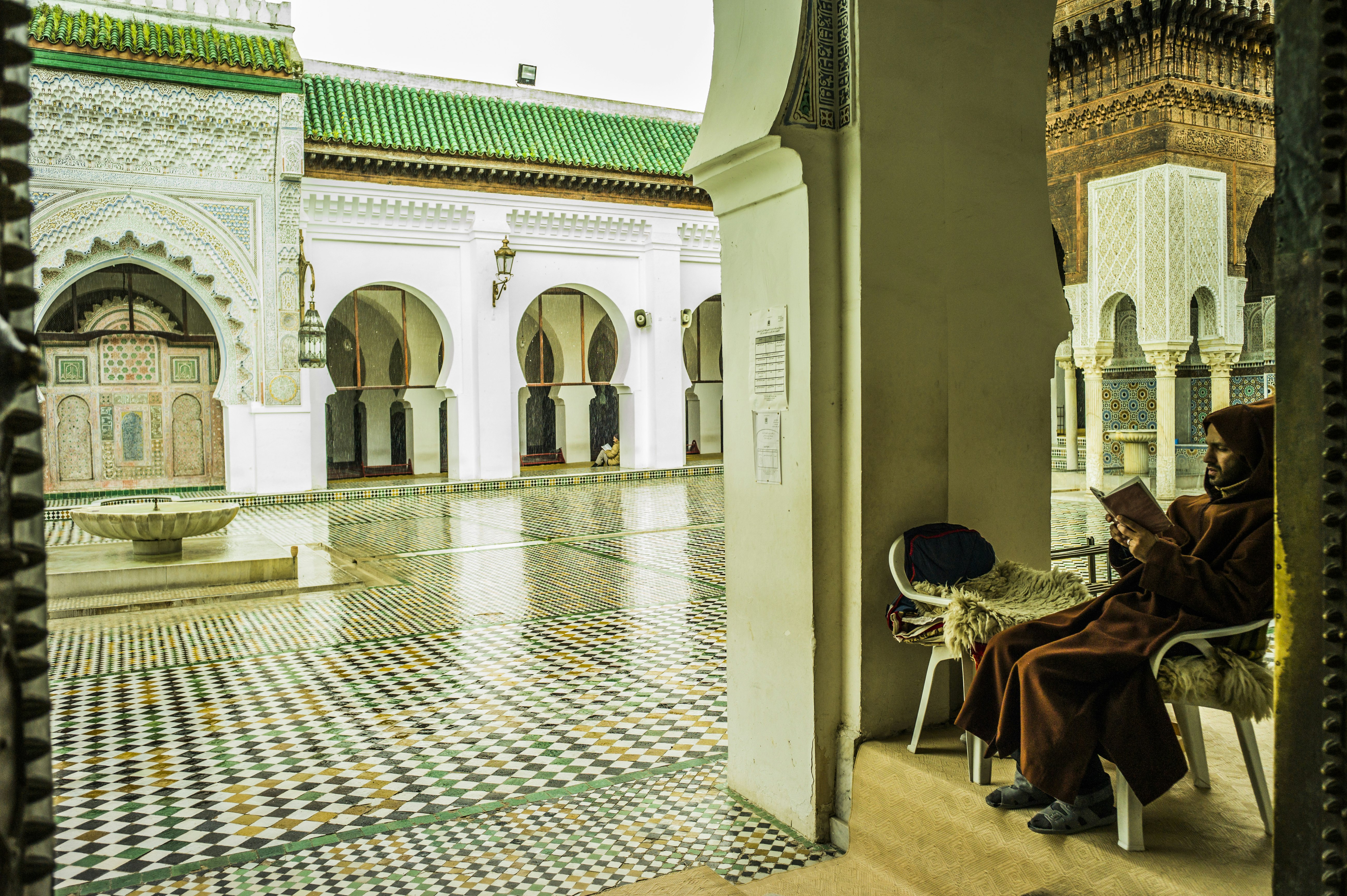 A man in brown robes reads a book tucked away in the ornate halls of the Kairaouine Mosque in Fes, where the floors are made up of gleaming mosaics, white arched walls and columns, and green tile roofs 