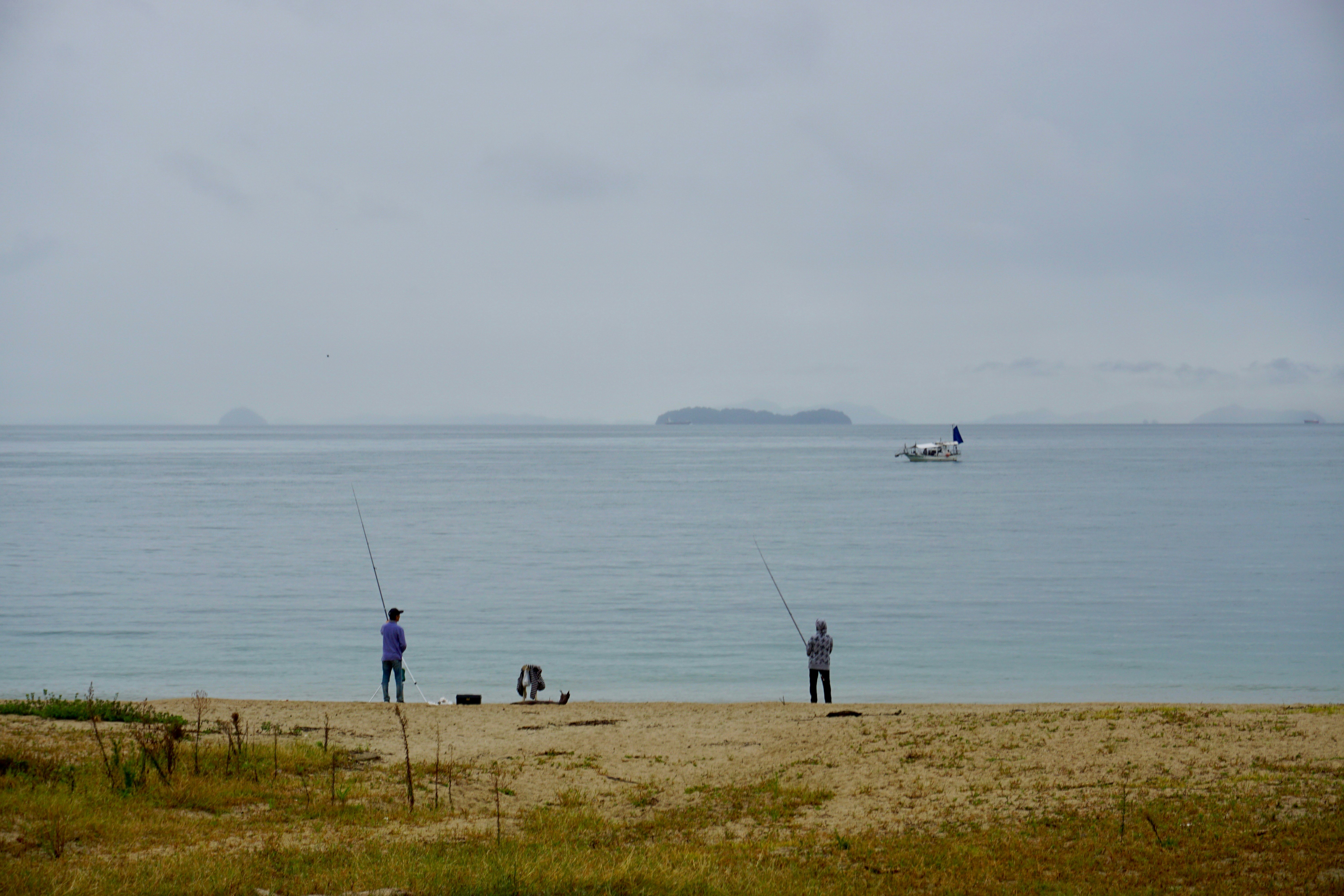 Two fishermen, one in a purple shirt and dark jeans and the other in a grey and black sweater and black slacks face the Seto Inland Sea with long fishing poles. In the distance is a white fishing boat with a navy blue sail, and even further in the distance are several small, mountainous islands almost obscured by the haze.