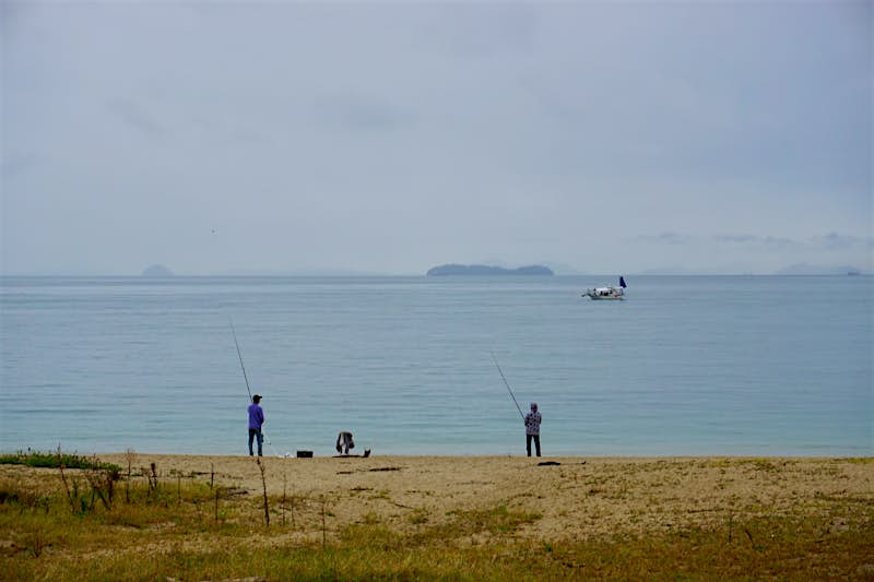 Kamikamagari.jpg Two fishermen, one in a purple shirt and dark jeans and the other in a grey and black sweater and black slacks face the Seto Inland Sea with long fishing poles. In the distance is a white fishing boat with a navy blue sail, and even further in the distance are several small, mountainous islands almost obscured by the haze.